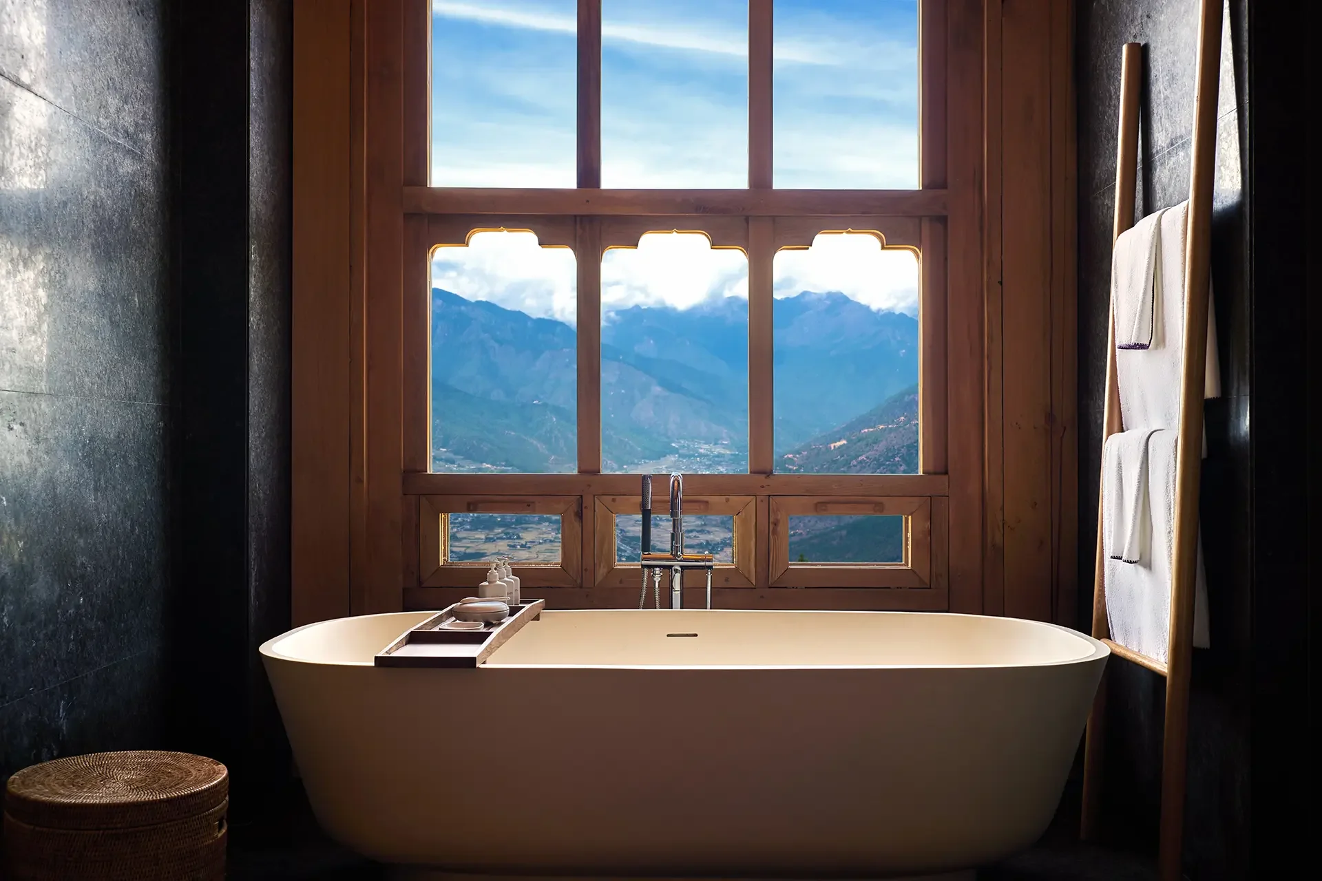 A bathroom with a freestanding bathtub placed in front of a wooden-framed window. The window offers a view of mountains and a partly cloudy sky. There are white towels hanging on a wooden towel rack on the right side, a small stool on the left, and bath supplies on a tray on the edge of the bathtub.