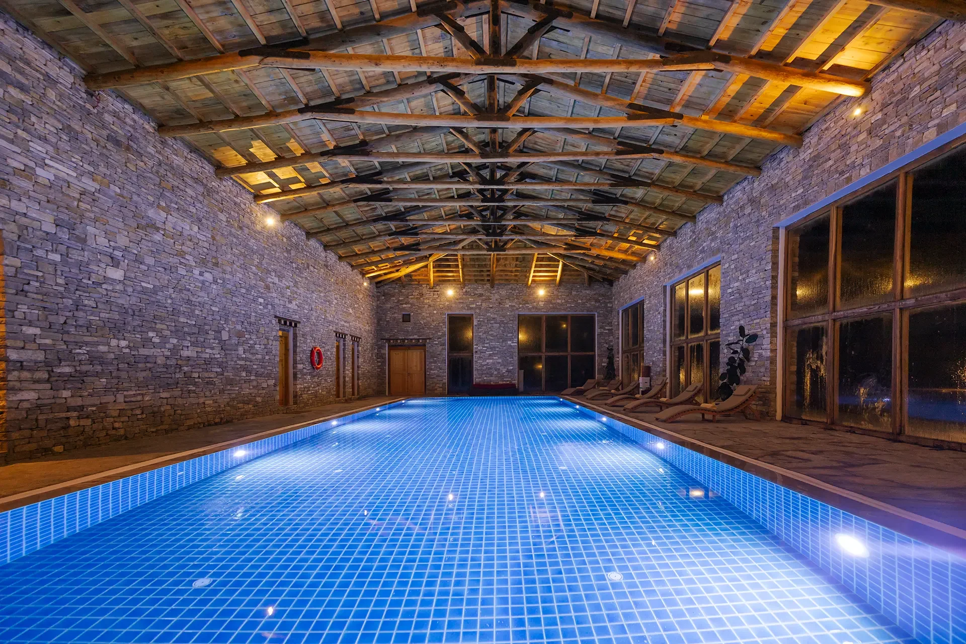 Indoor swimming pool with blue tiling, surrounded by stone brick walls, large windows, and wooden ceiling beams, illuminated by warm lighting.