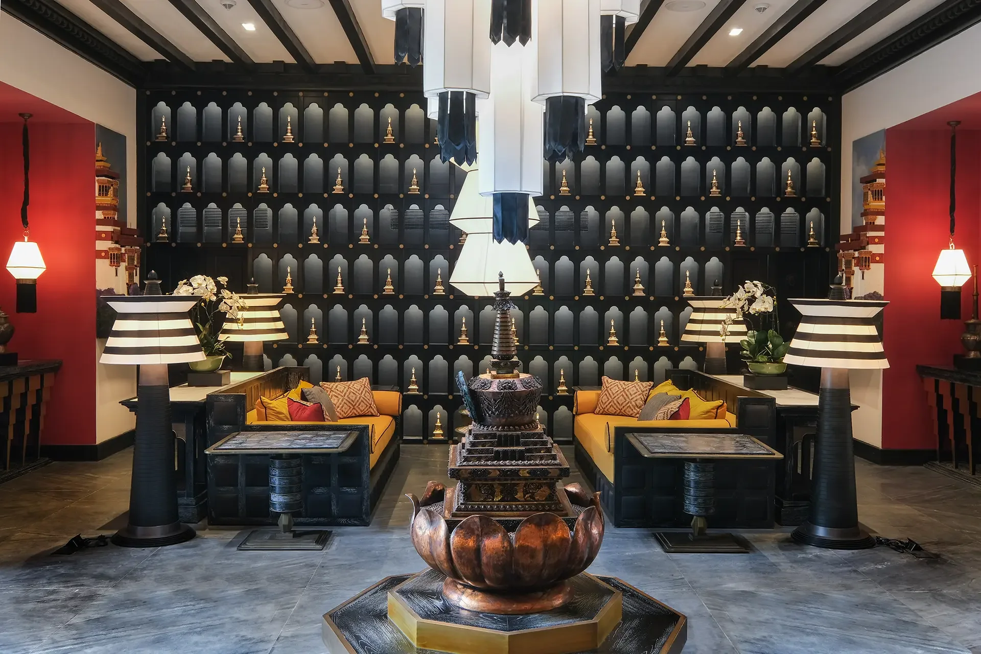 Interior of a room with a black wall decorated with small golden Buddha statues, surrounded by red and white walls, featuring yellow sofas with patterned cushions, black tables, and decorative lamps, with a central fountain.