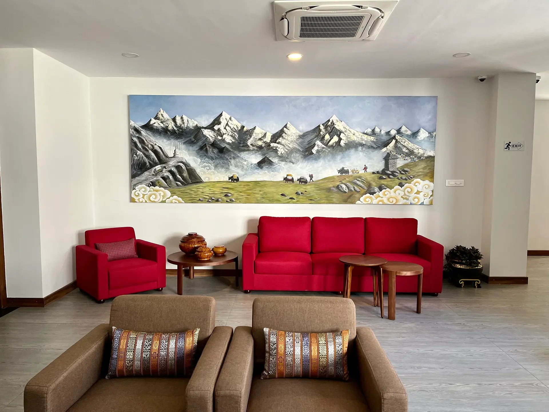 Indoor lounge area with beige and red seating, a wall painting of snow-capped mountains, giraffes, oxen pulling carts, and prayer flags, and a small table with decorative items.