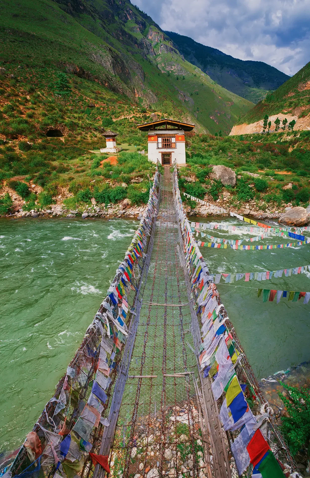 A suspension bridge decorated with colorful prayer flags spans across a river in a lush green valley surrounded by mountains, leading to a traditional building with a sloped roof.