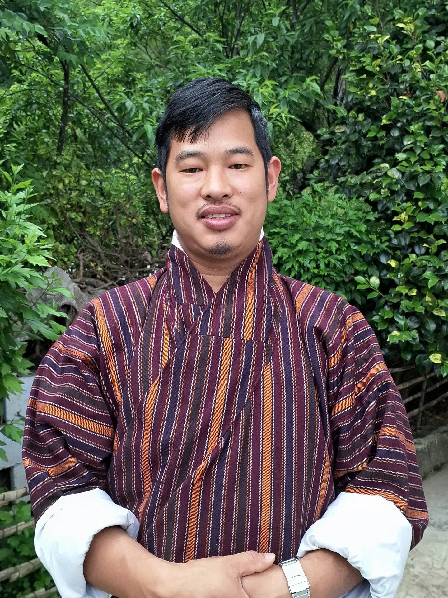 A man wearing a traditional striped garment standing outdoors in front of green foliage.