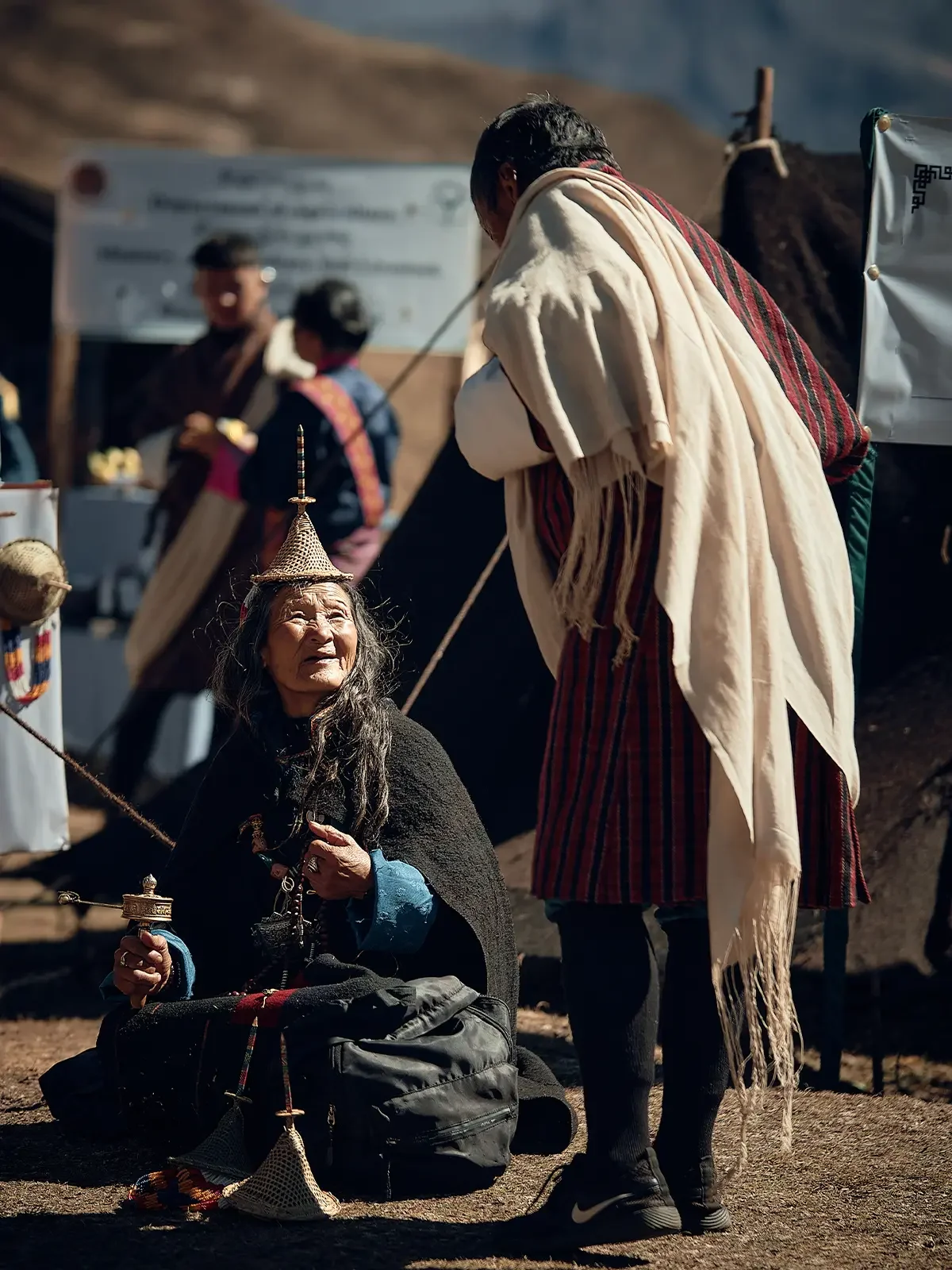 An elderly woman is sitting on the ground, holding a prayer staff, wearing traditional clothing and a small, cone-shaped hat. A man standing in front of her, dressed in traditional attire, is bending forward, engaging with her. In the background, the