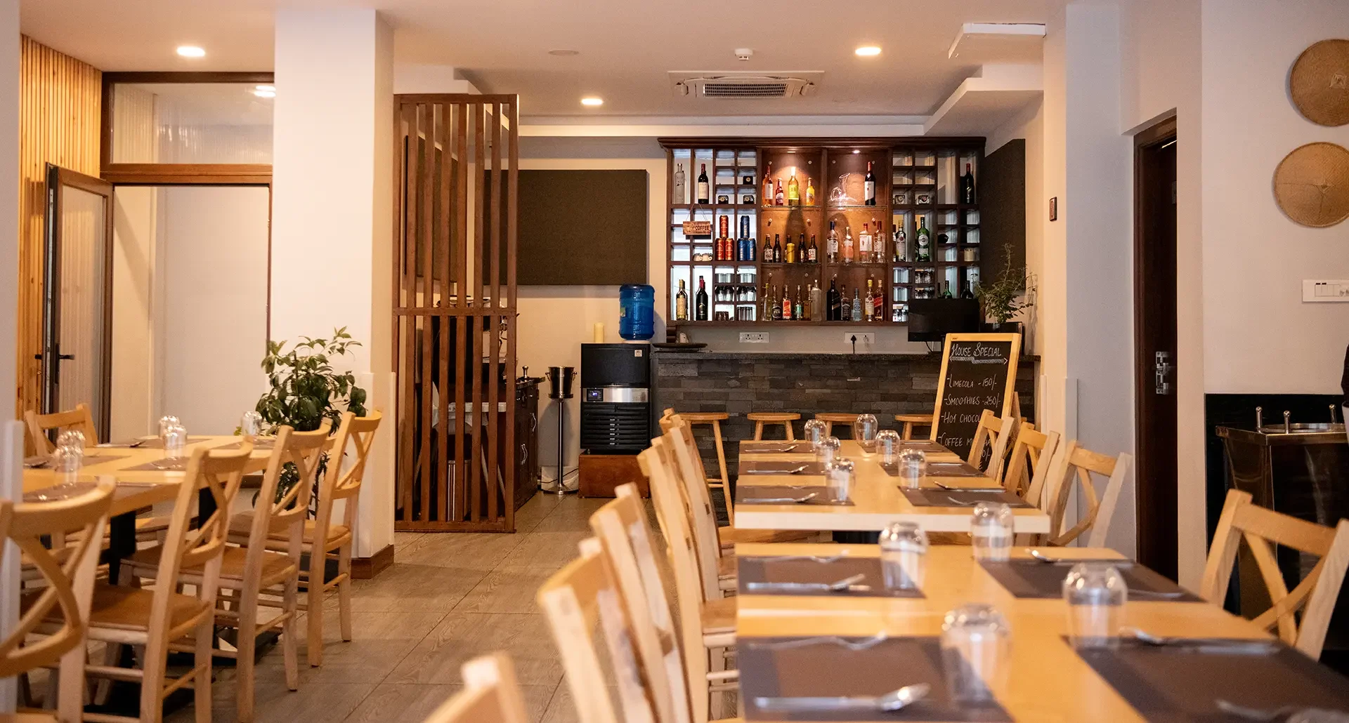 Interior of a restaurant with wooden tables and chairs, a bar with bottles, a chalkboard menu, and minimal decor.