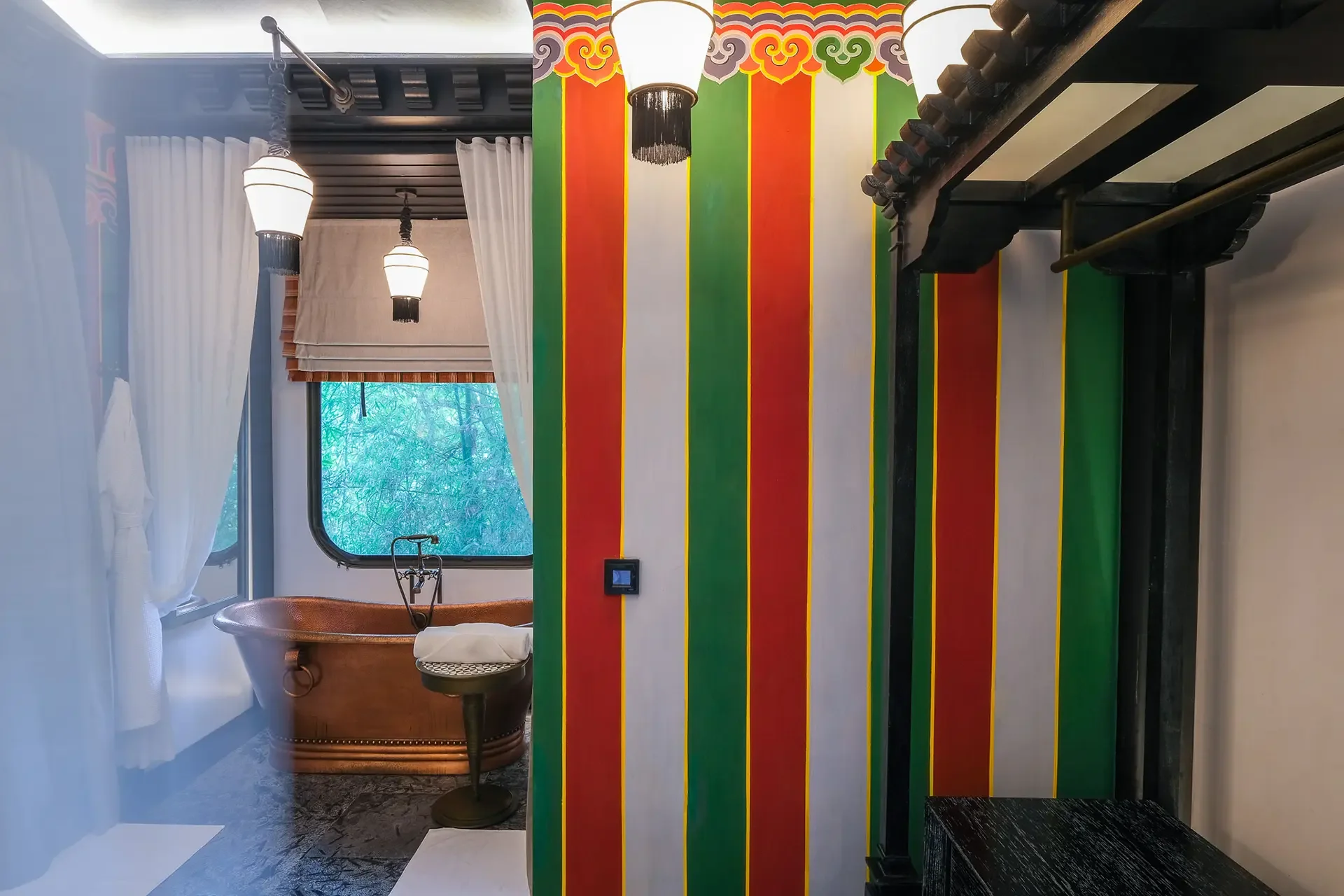 Colorful striped wall with red, green, white, purple, and yellow stripes. Black soundproofing and furniture in the room include a black bench and a copper bathtub. Two hanging lamps with white shades, large window with white curtains, and a small table with a metal stand are also visible.