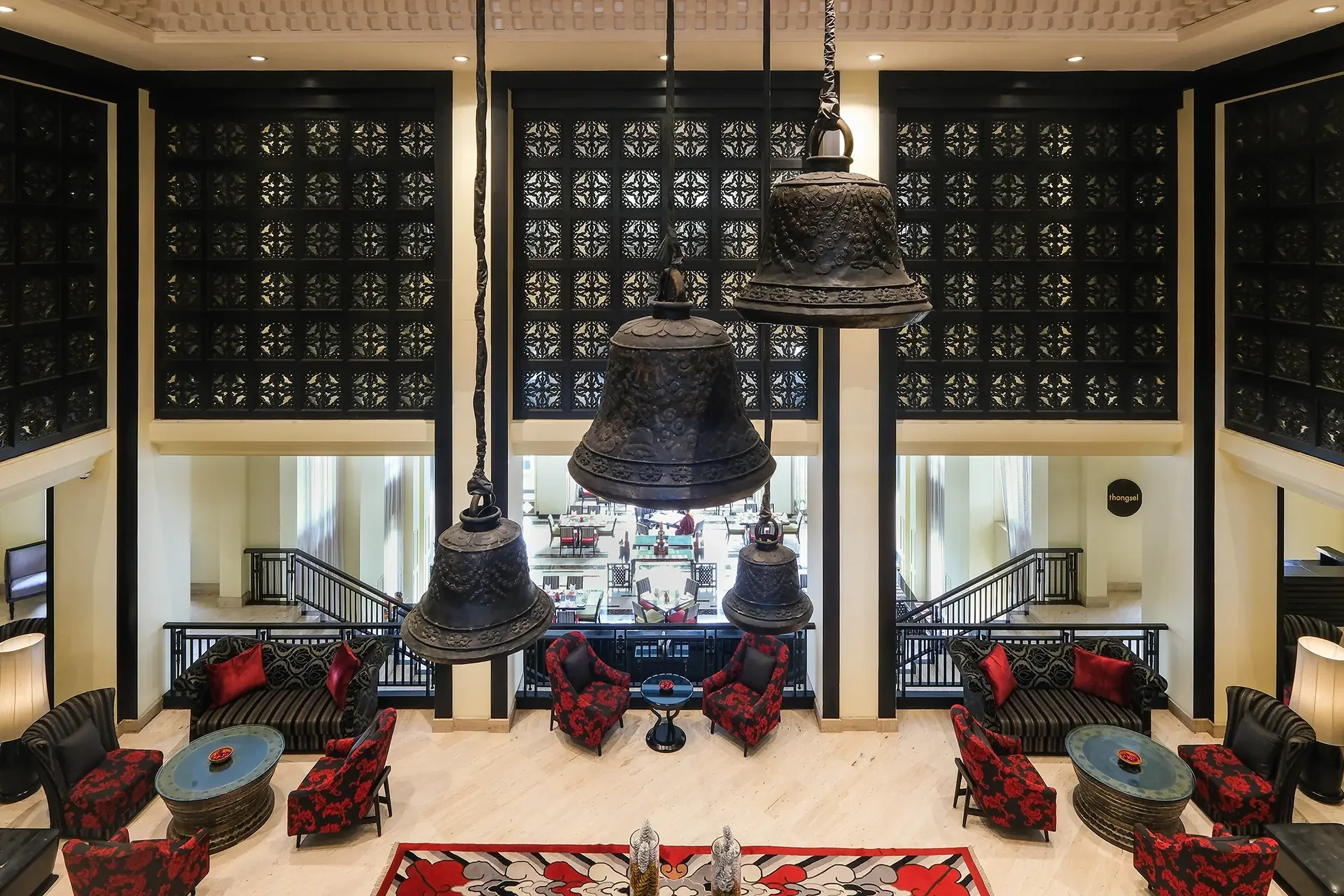 Interior of a hotel lobby with large bronze bells hanging from the ceiling, surrounded by black and red chairs, tables, and decorative panels on the walls.
