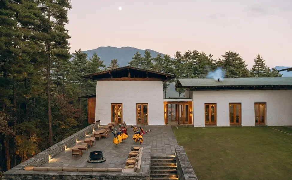 An outdoor patio area attached to a modern house surrounded by trees, with a mountainous background and a clear sky. There are people dressed in colorful traditional clothing performing a dance or celebration.