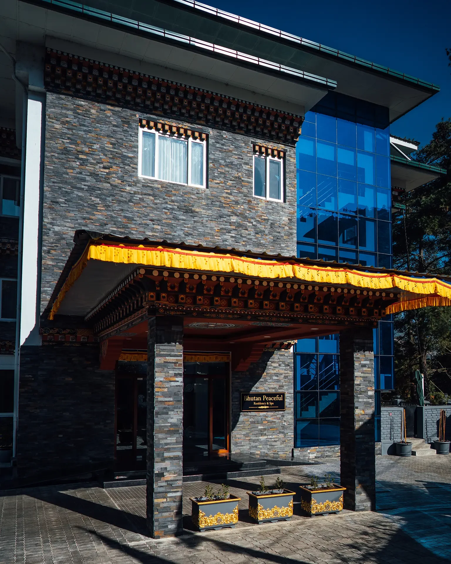 Modern hotel building with stone facade, featuring a sign that reads 'Bhutan Peaceful Residency & Spa,' decorative gold planters in front, and traditional Bhutanese architectural elements under the roof canopy.