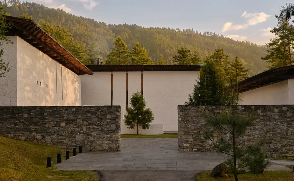 A modern building with white walls and a stone wall front, surrounded by greenery and trees, with a mountain in the background.