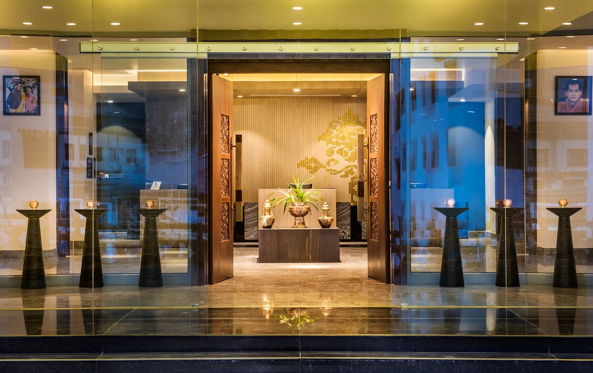 Hotel lobby entrance with glass doors, black pedestals with lit candles, decorative vases, and a large potted plant at the center of an elegant reception area. Framed portraits hang on the wall, and a golden Asian-inspired artwork decorates the back wall.