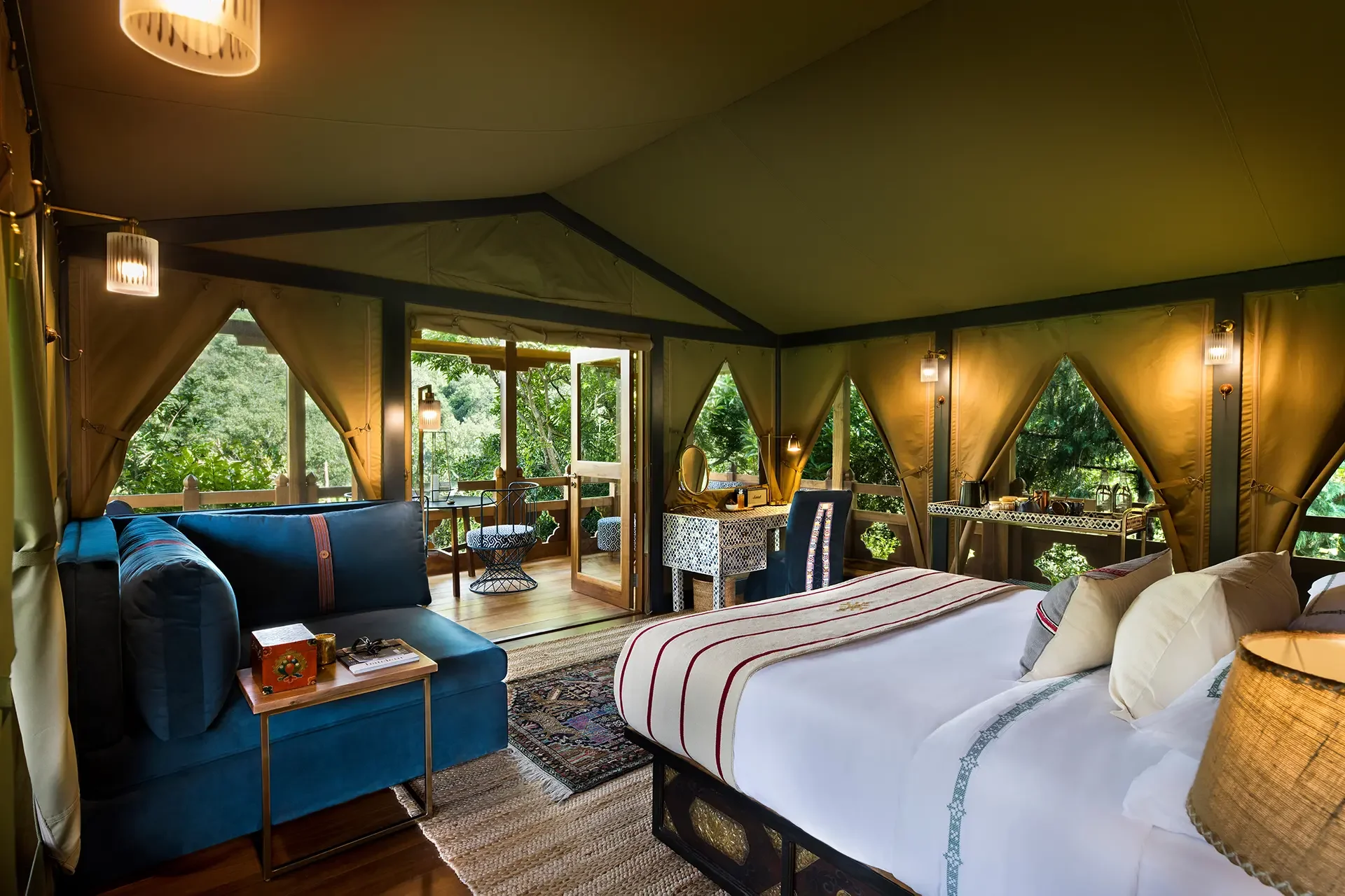 Interior of a cozy glamping tent with large windows revealing green trees outside. The room has a bed with white linens, pillows, a blue velvet sofa, a small wooden side table, and a dresser with a mirror. Warm lighting fixtures hang from the ceiling, and a balcony with chairs is visible through the open door.