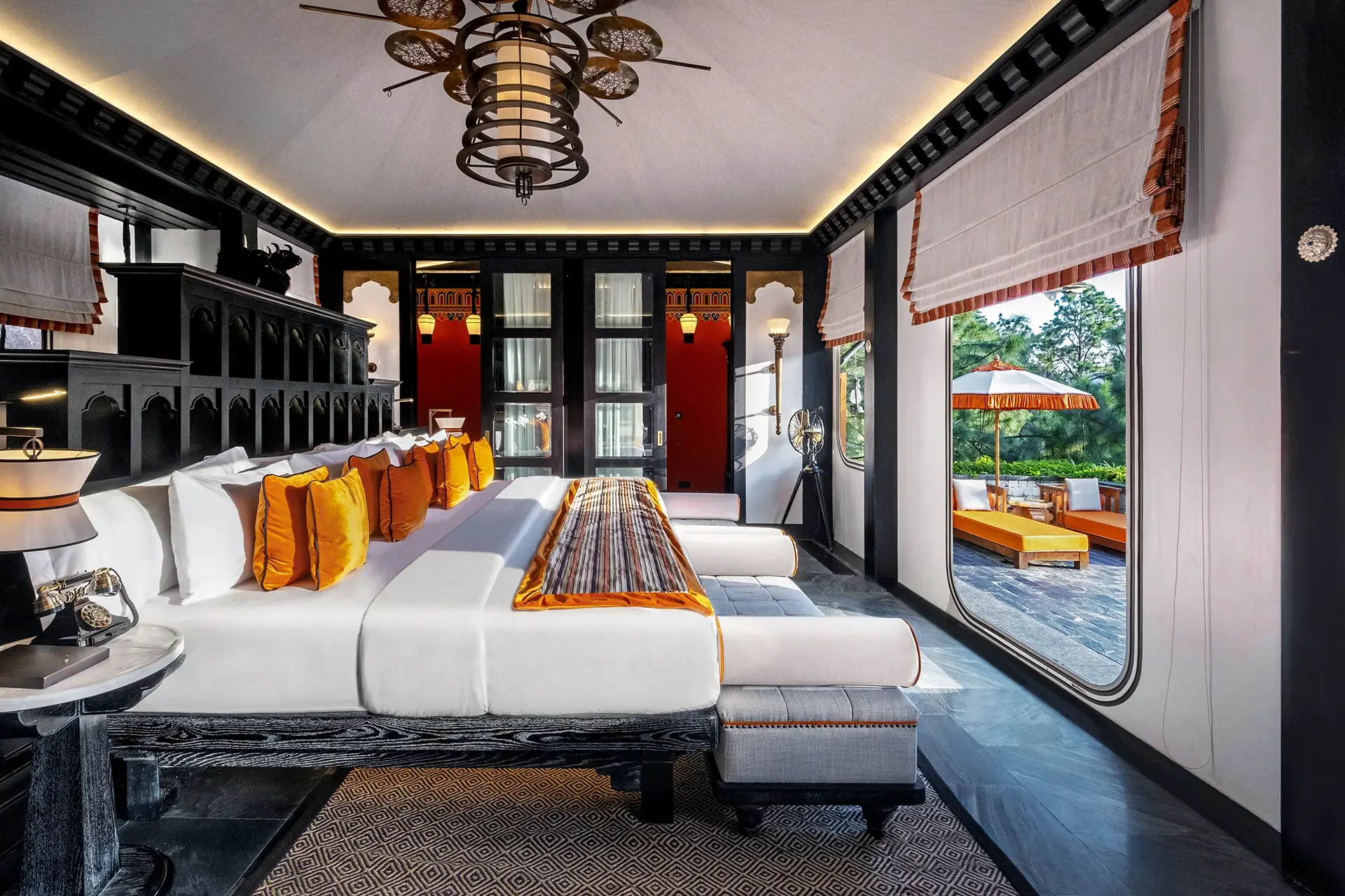Elegant bedroom with a white bed and orange accent pillows, black wooden headboard, decorative ceiling light, and a sliding glass door leading to a patio with outdoor seating and green trees.