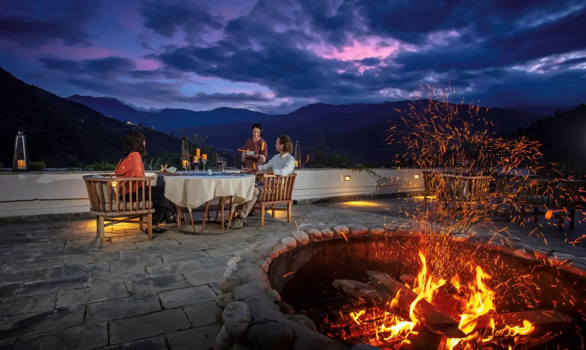 A romantic outdoor dinner scene at dusk with three people, two women and a man, sitting at a table on a mountain terrace. A server is taking their order. A fire pit with sparks burning is in the foreground. The sky is dark with clouds and purple hues, and mountains are visible in the background.