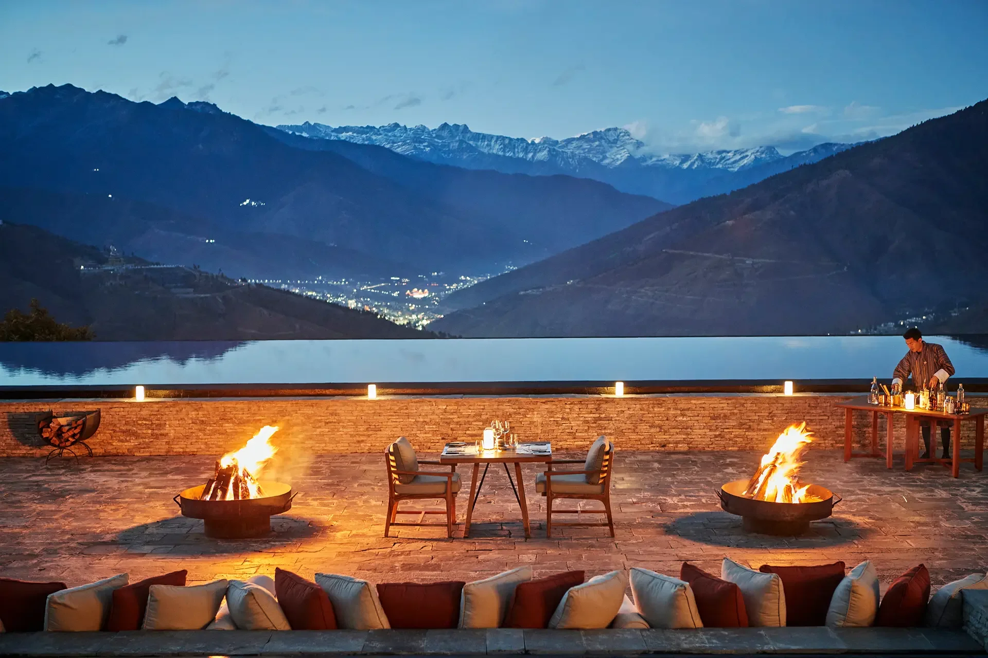An outdoor patio with a firepit, chairs, and a table with candles overlooking a mountain landscape at dusk, with a person preparing drinks.