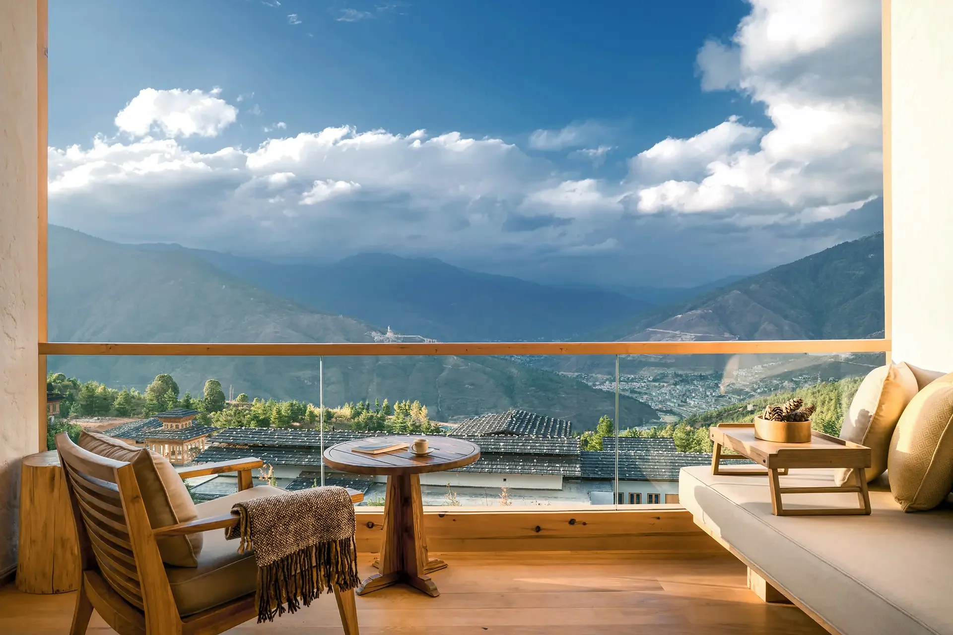A cozy balcony with wooden furniture including a round table with a coffee cup, a cushioned armchair with a blanket, and a sofa with pillows. The balcony has glass railing overlooking a mountain landscape with clouds, green hills, and rooftops of houses below.