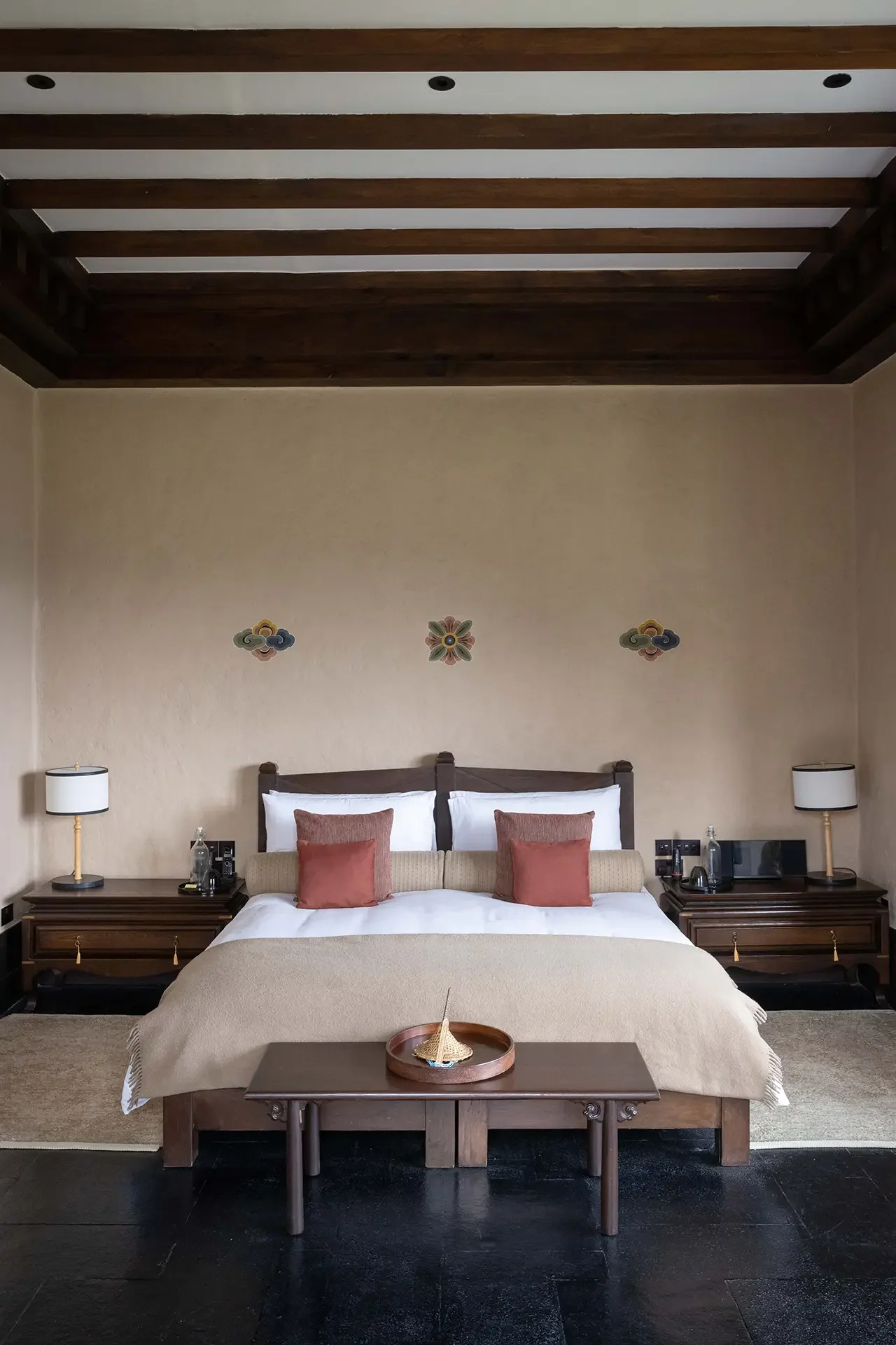 A bedroom with a large bed, beige bedding, and four peach-colored pillows. Wooden nightstands with lamps and water bottles are on each side of the bed. The wall behind has three decorative wall hangings with cloud and floral motifs. The ceiling has wooden beams and recessed lighting.