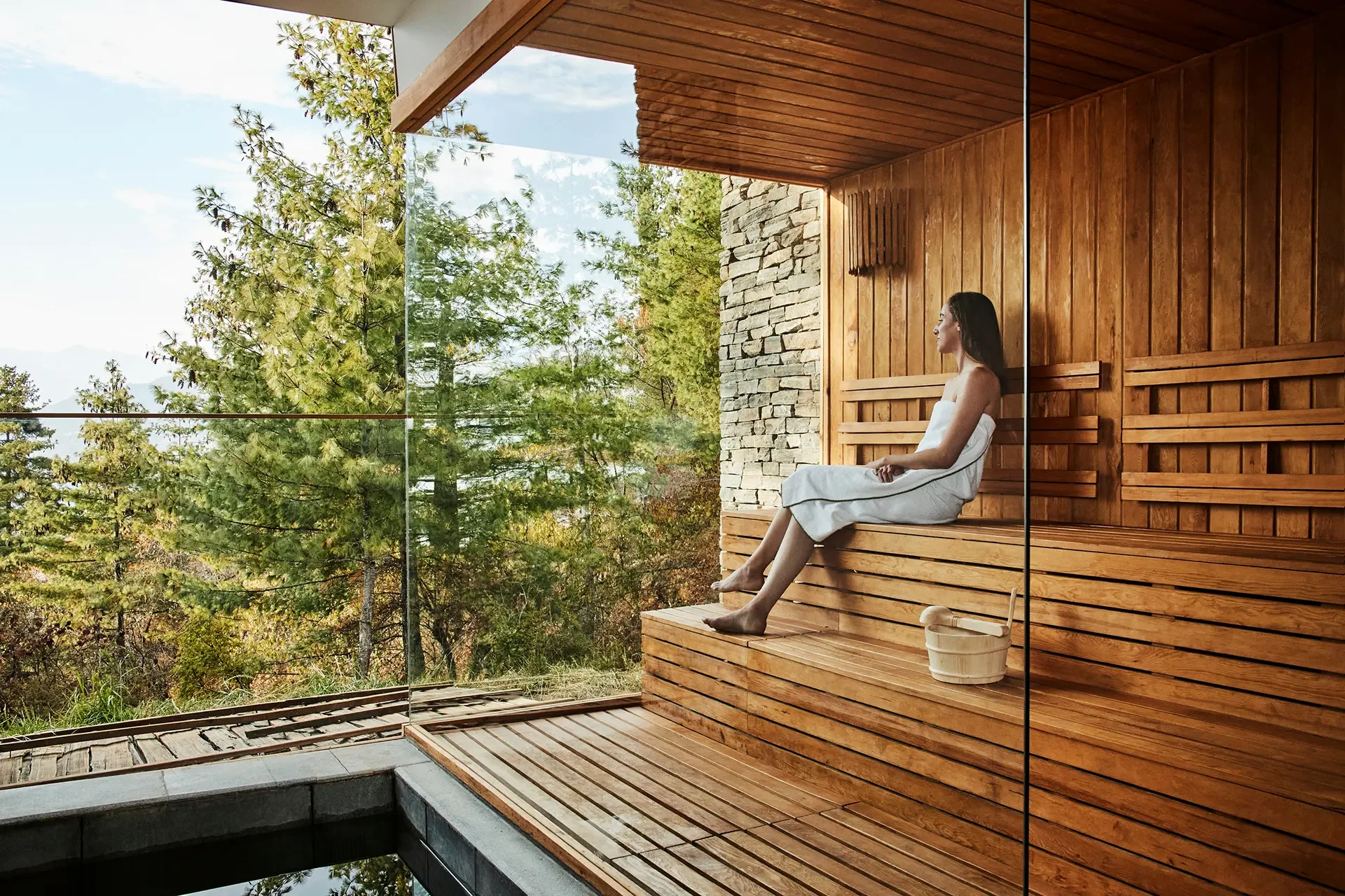 A woman sitting wrapped in a towel on a wooden bench in a sauna, with a wooden bucket and ladle nearby, looking out through a glass wall at lush green trees and a blue sky.