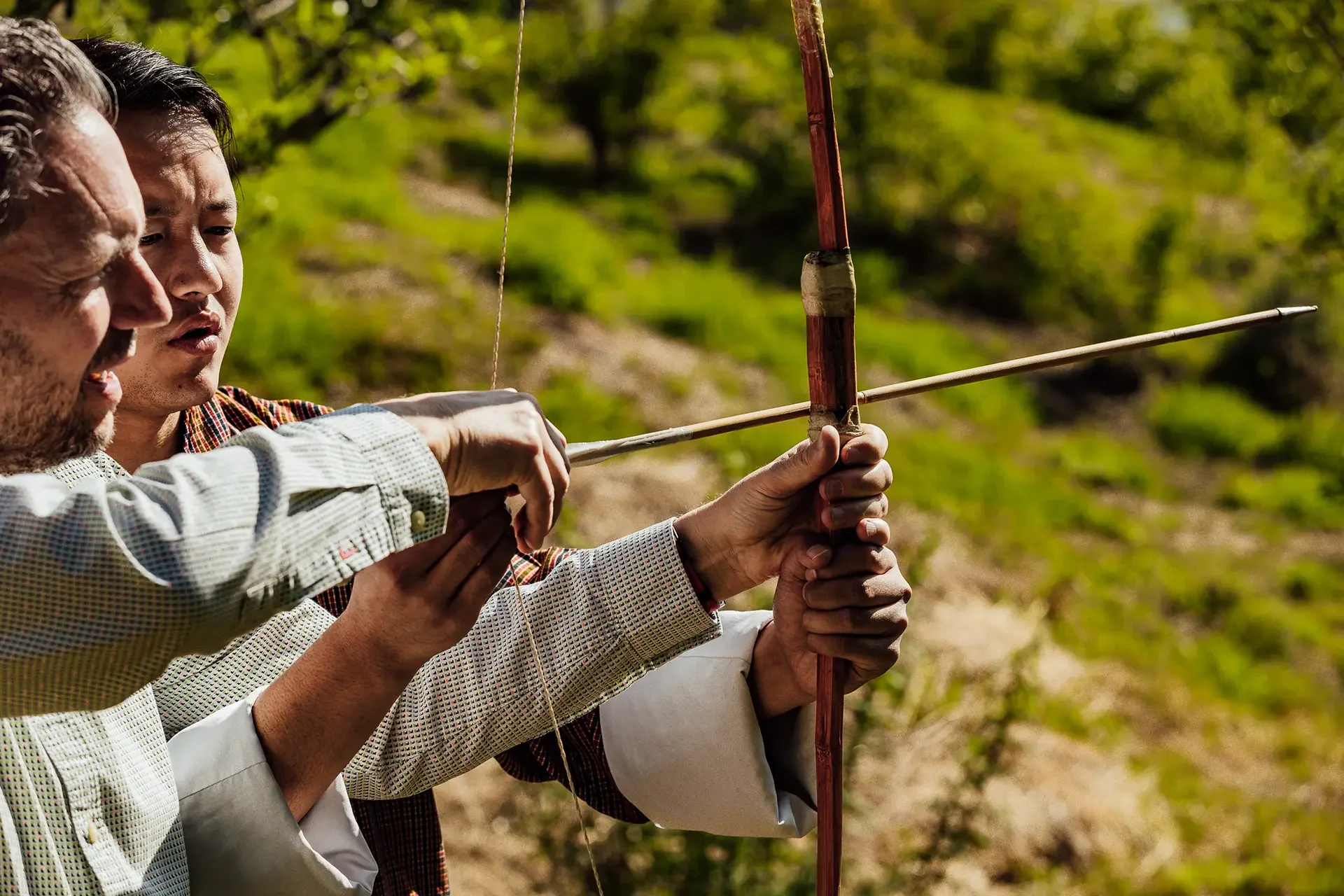 Two men holding a traditional bow and arrow in a green outdoor setting.