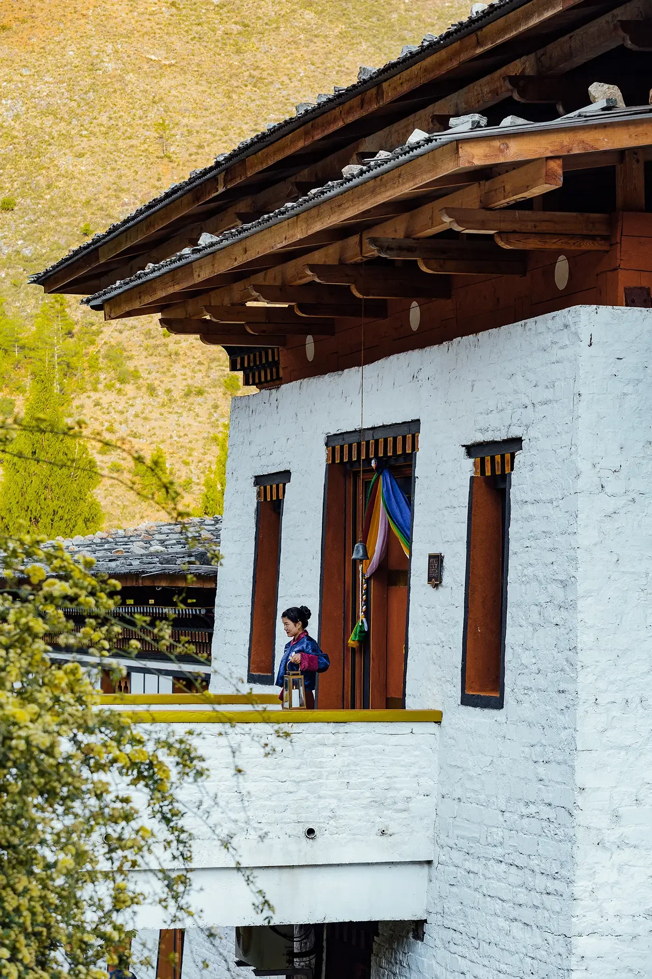 A woman dressed in traditional attire standing outside a white and wooden building with a bell and colorful fabric banner hanging near the door, surrounded by trees and mountain landscape.