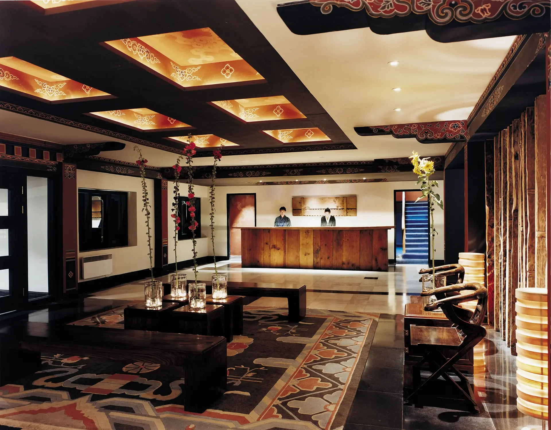 Lobby area with a wooden reception desk, decorative ceiling panels, floral plants hanging, framed artwork, patterned rug, chairs, and a staircase in the background.