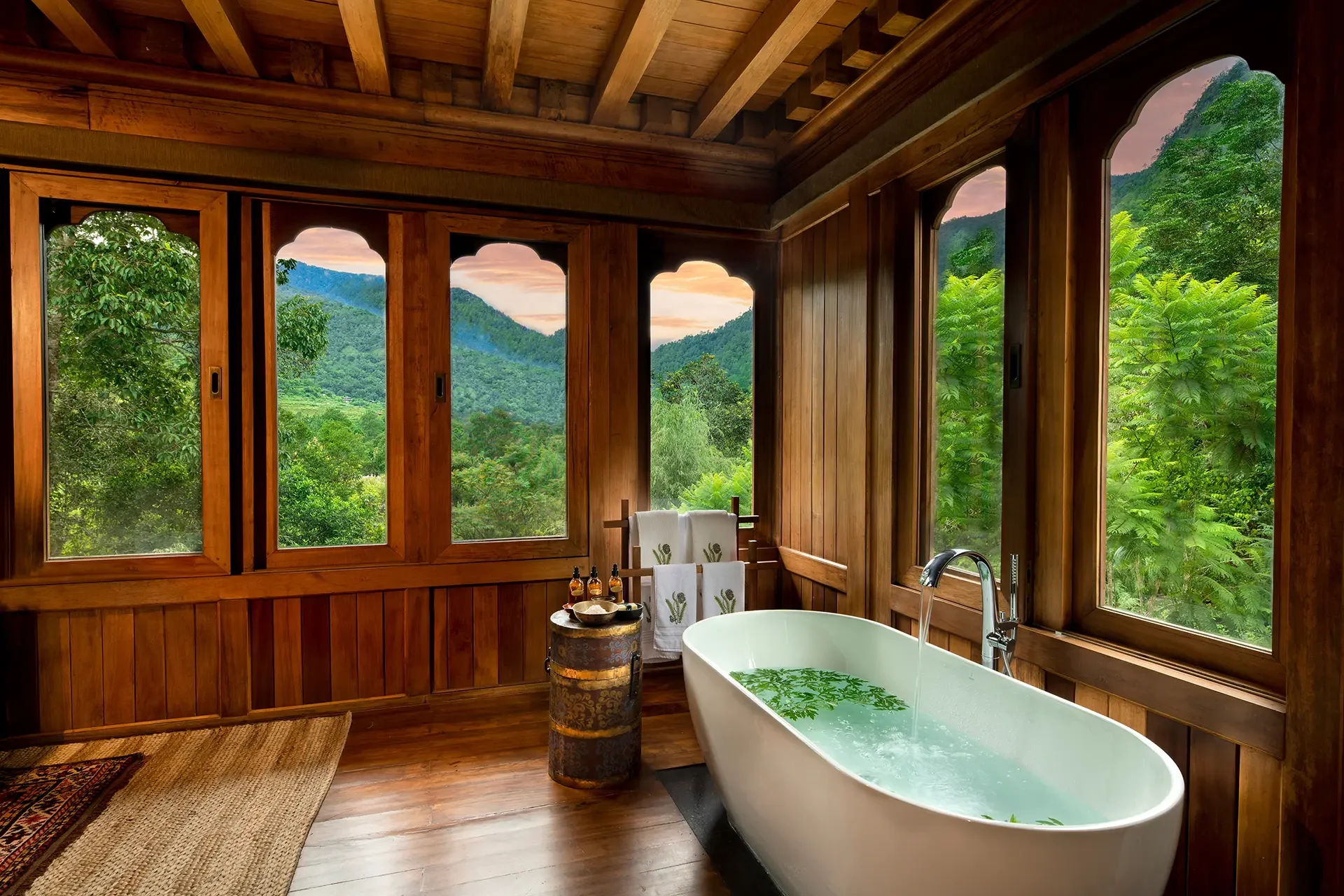 A rustic bathroom with wooden walls and ceiling featuring large windows showing a lush green mountain landscape. Inside, there is a freestanding bathtub filled with water and floating green leaves, with a small table holding bath products nearby. Towels hang on a towel rack, and a rug is on the wooden floor.