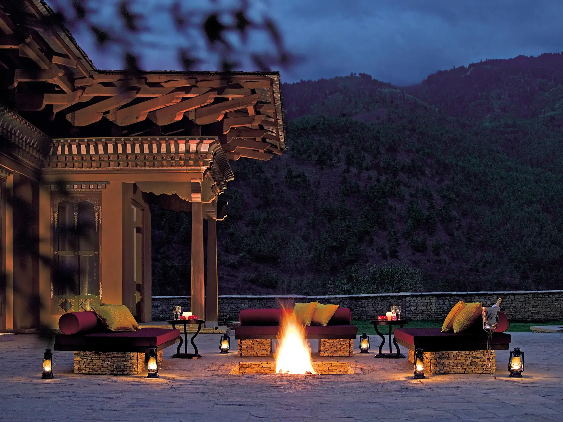Outdoor seating area with a fire pit, surrounded by benches and small tables, set against a mountain backdrop at dusk.