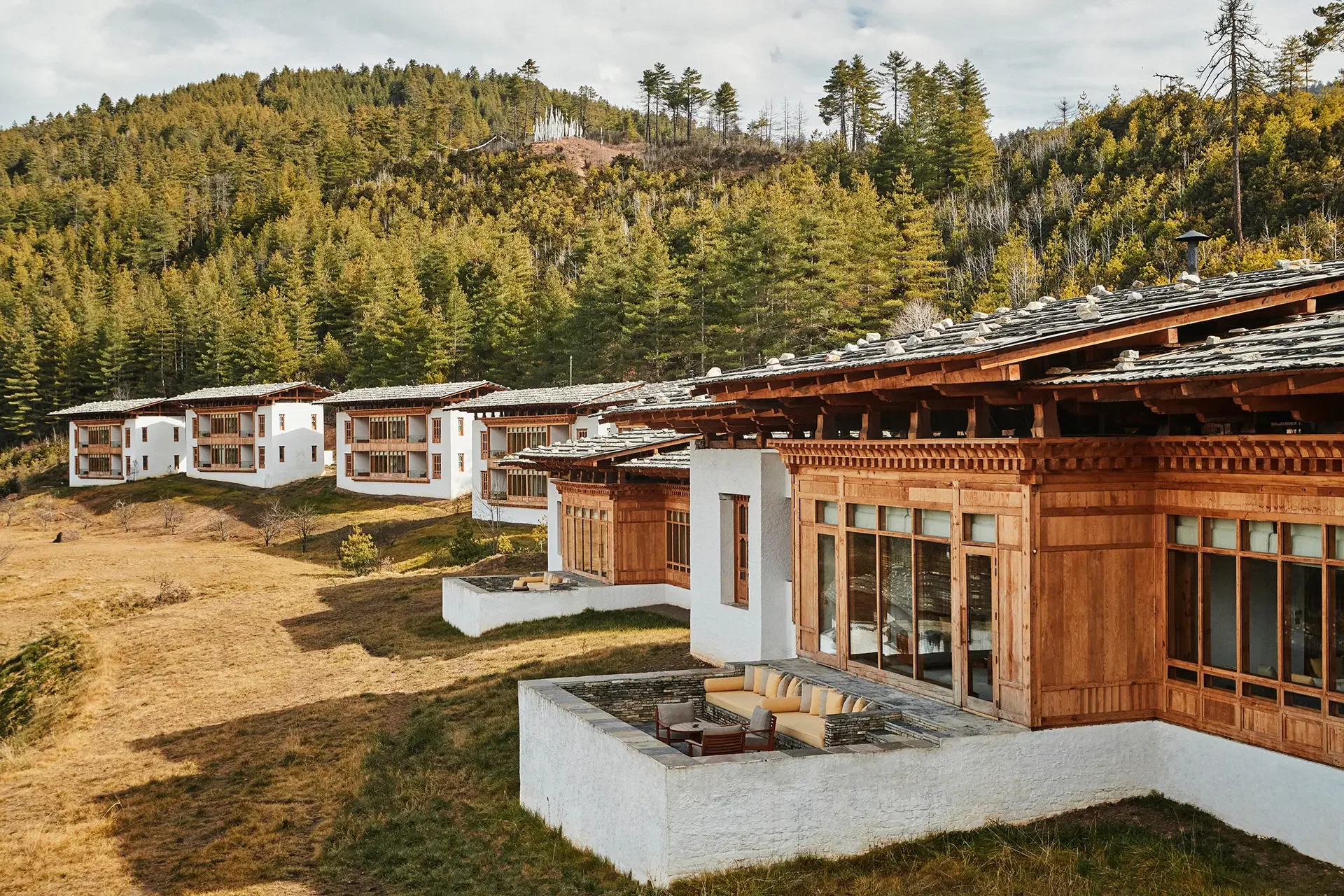 Multi-story residential homes with wooden balconies and white exterior walls, set against a hillside covered with pine trees, under a cloudy sky.