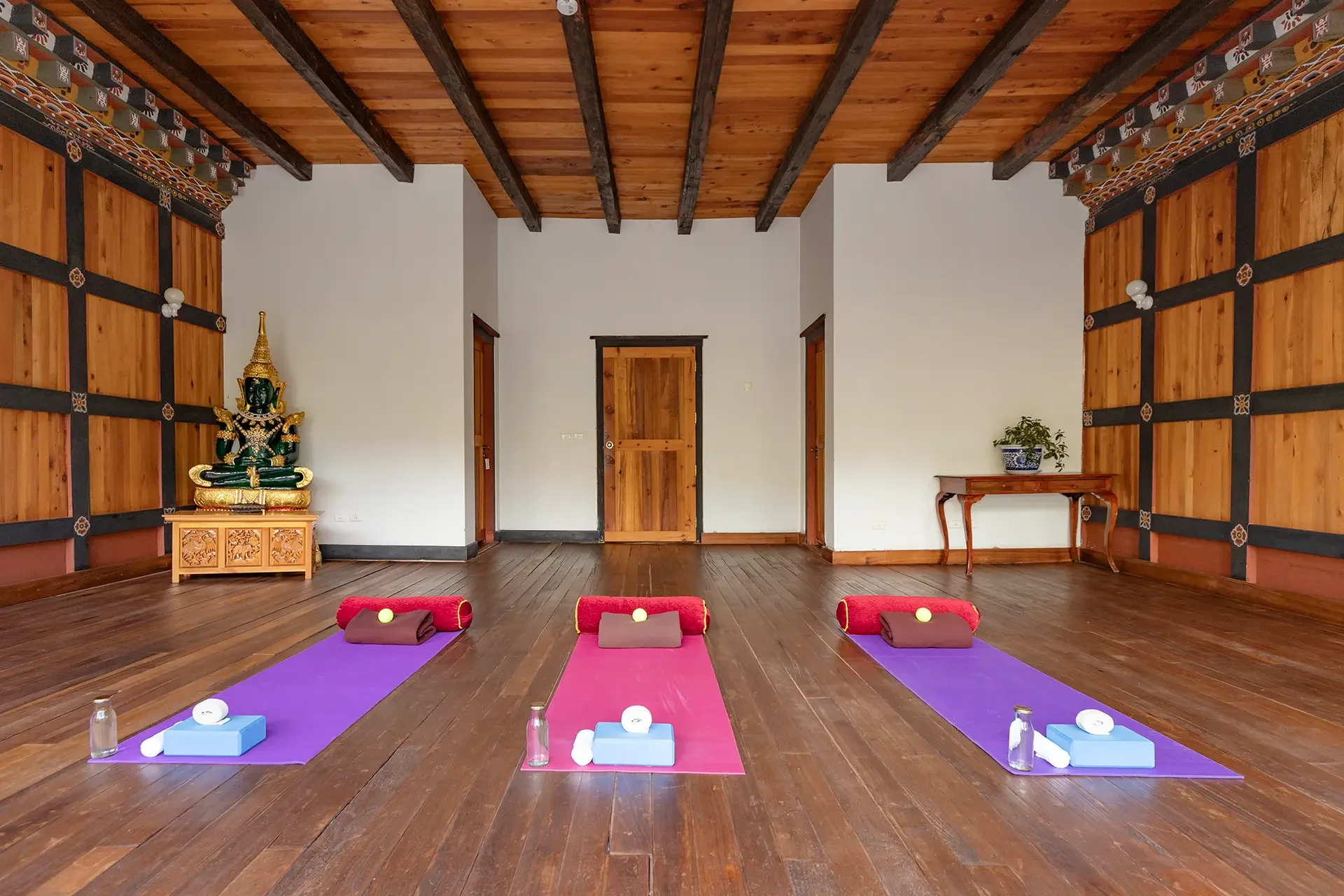 An indoor yoga studio with wooden floors and walls, three yoga mats in purple and pink colors with towels and cushions, water bottles, and yoga balls, a wooden table with a potted plant, wooden doors, and a statue of a deity in gold and green on a small carved wooden stand.