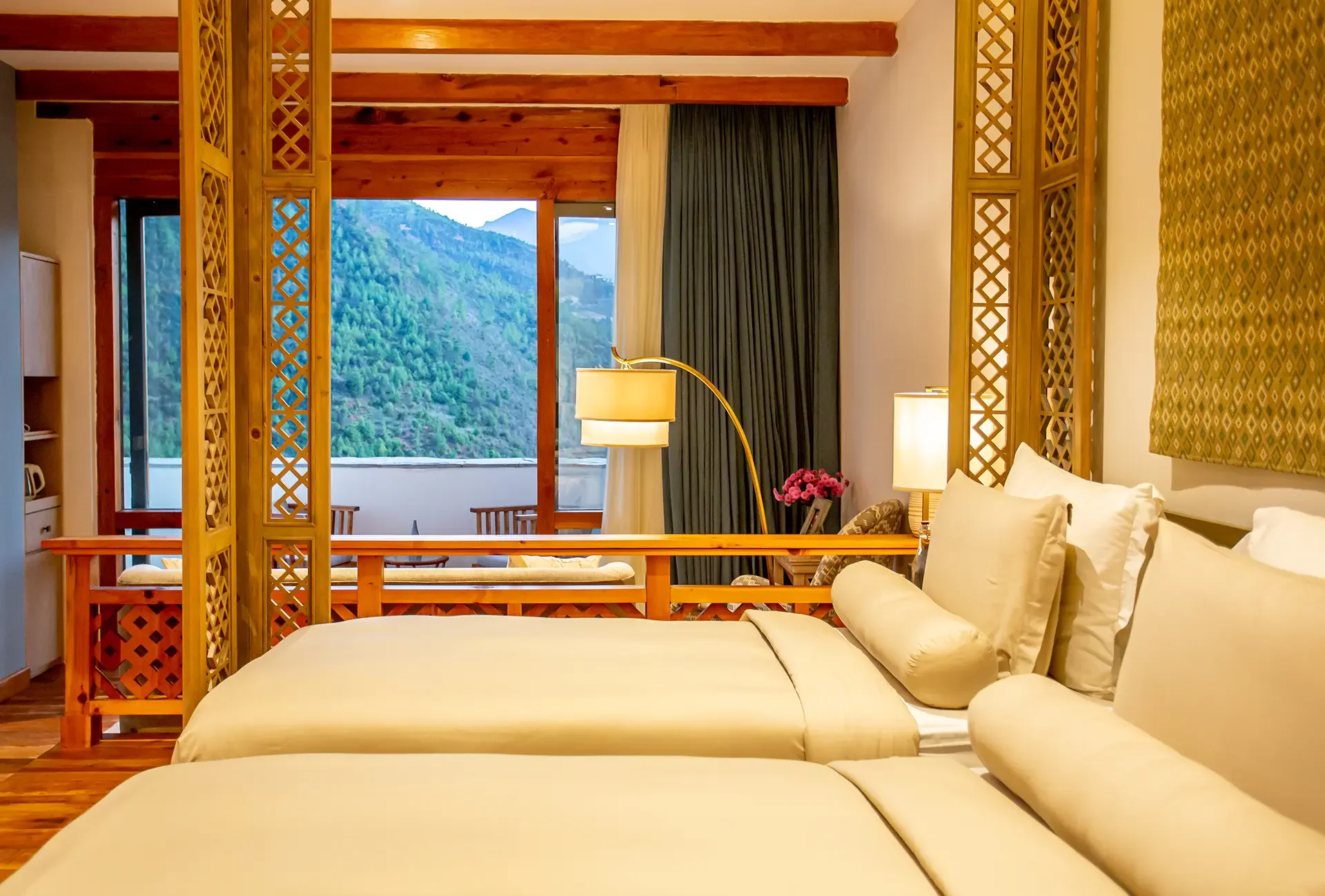A hotel room with two neatly made beds, wooden decor, large window with mountain views, indoor lamps, and patterned curtains.