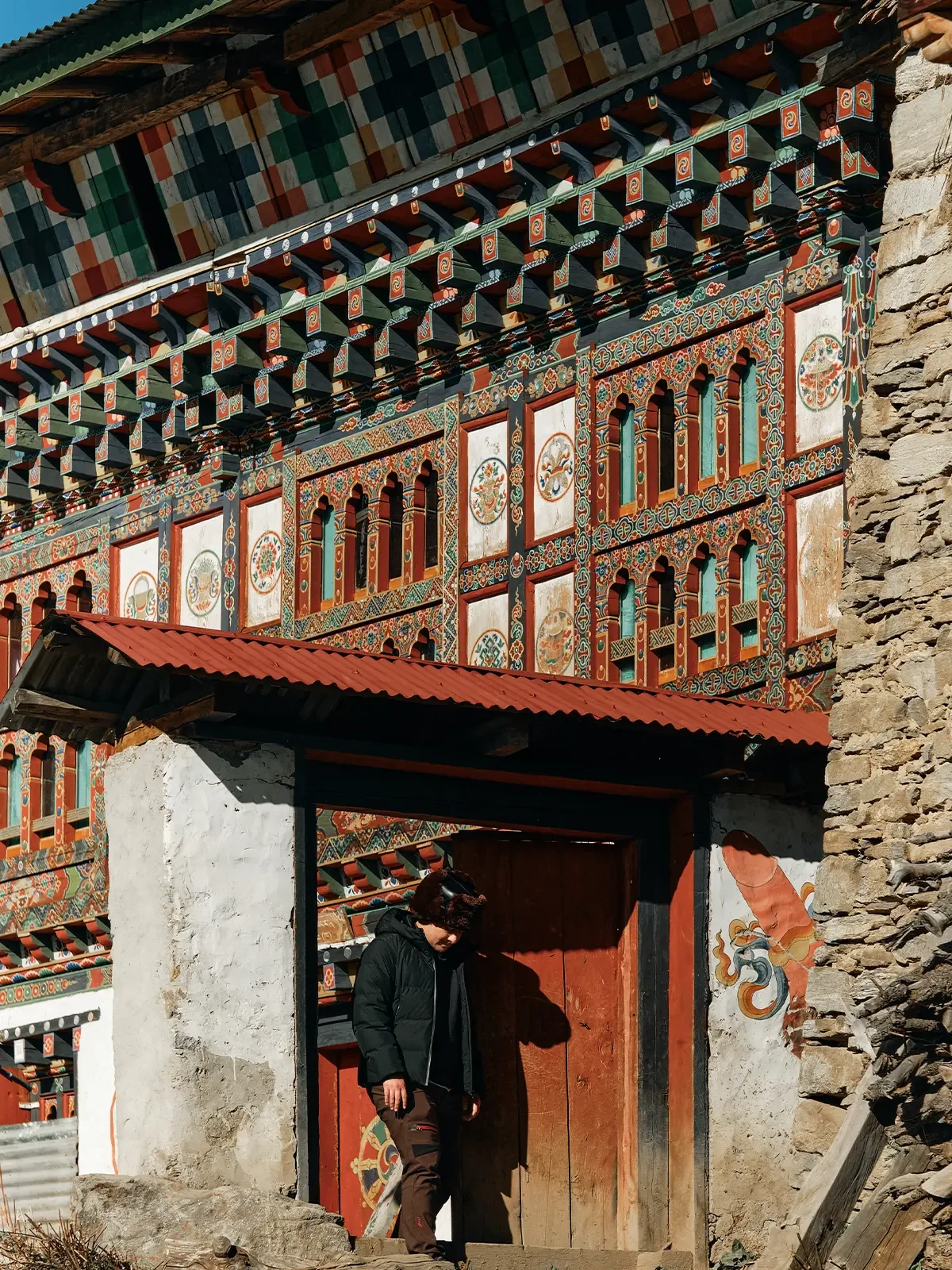 A person standing in front of a traditional building with ornate, colorful wooden decorations, a stone wall, and a red corrugated metal roof.