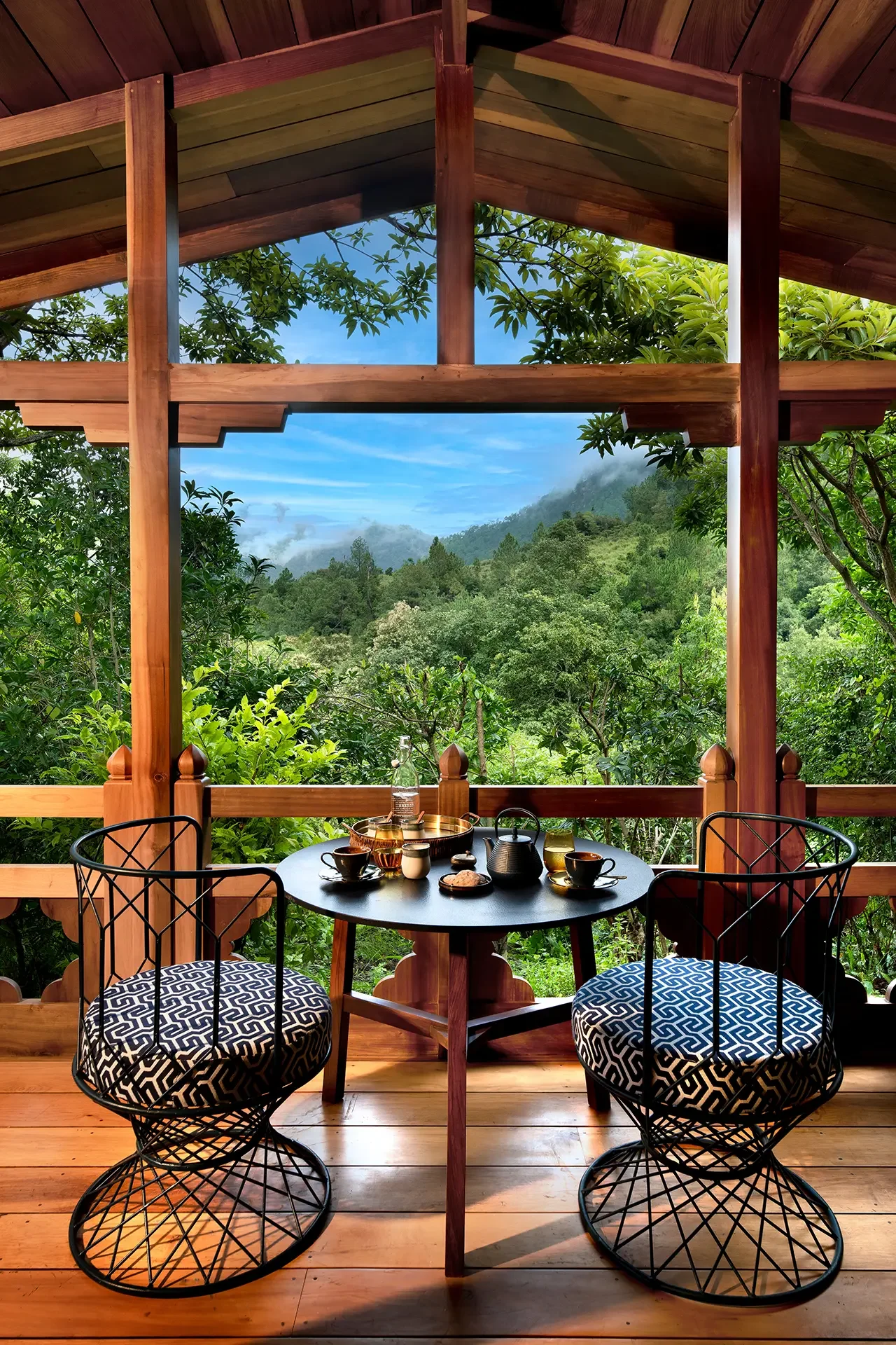 A cozy outdoor dining area on a wooden deck with a view of lush green trees and mountains in the background. There is a round black table set with cups, teapots, and snacks, surrounded by black wire chairs with geometric-patterned cushions.
