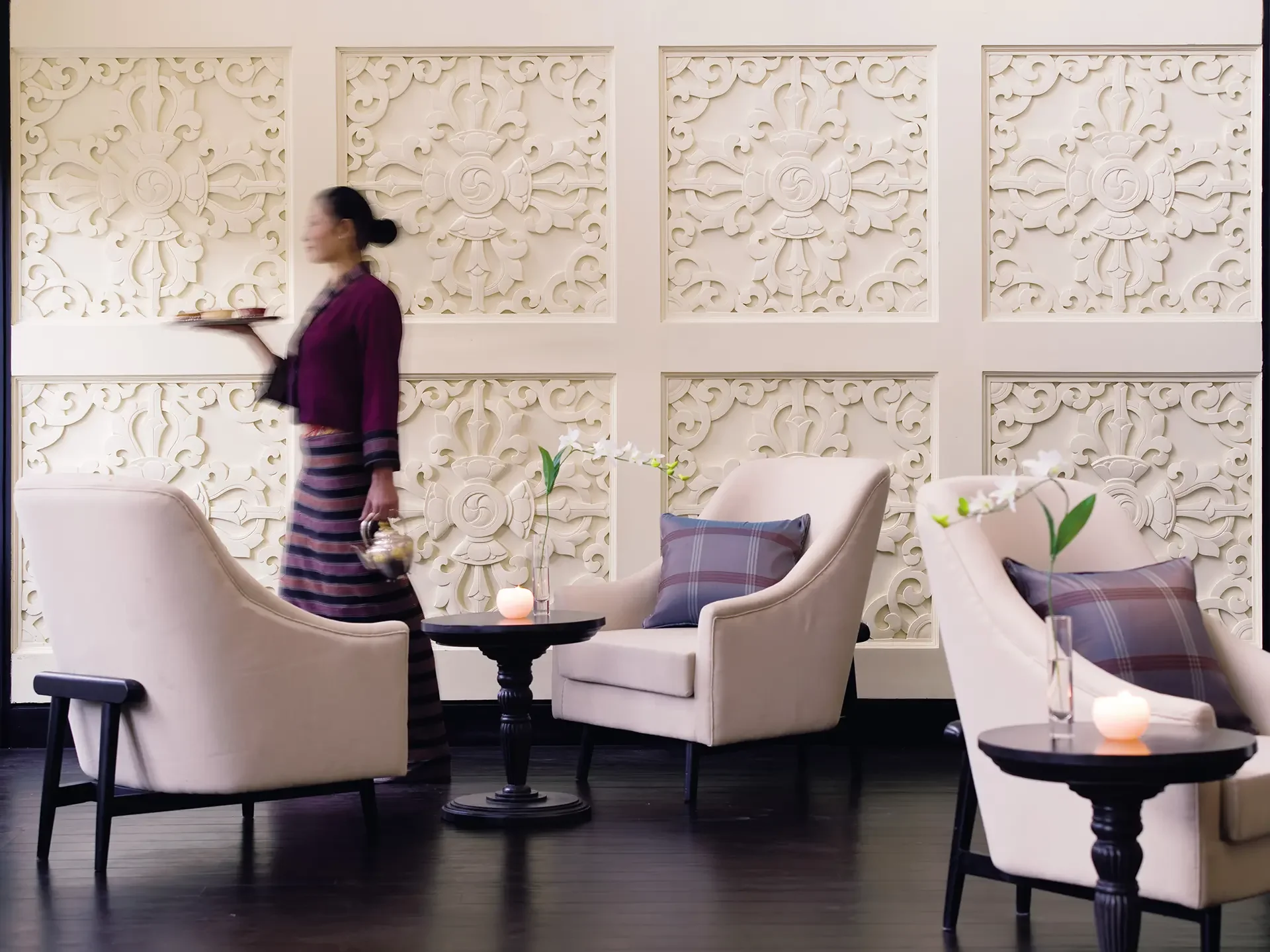 Interior of a modern lounge or restaurant with white upholstered chairs, small black tables, and decorative wall panels with an intricate pattern. A woman dressed in a purple and striped outfit is walking while carrying a tray. There are small candles with white flowers on the tables.