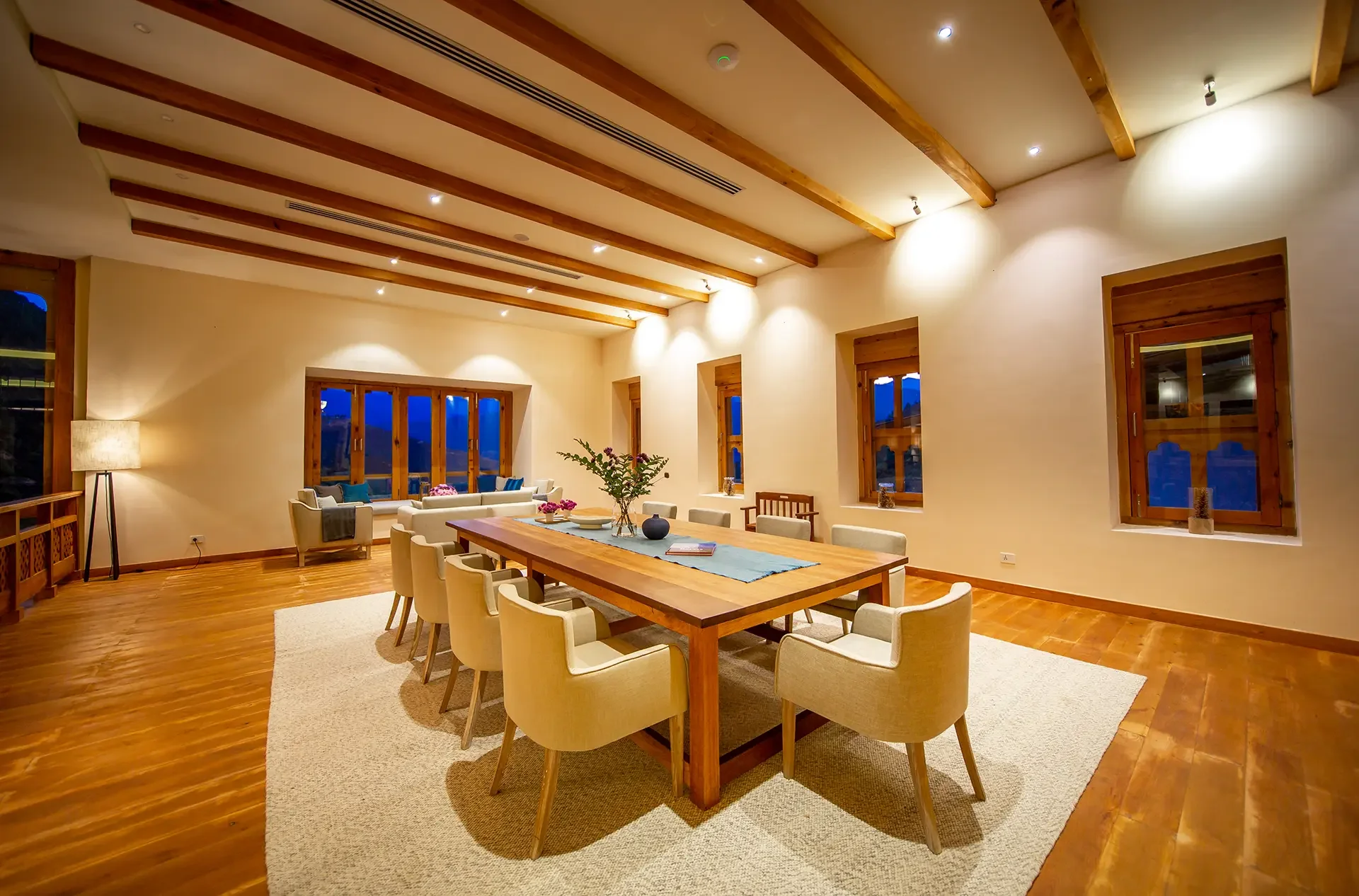 A spacious dining room with wooden beams on the ceiling, beige walls, and large windows showing a nighttime outdoor view. There is a large wooden dining table surrounded by beige chairs, a beige rug, and a centerpiece with flowers. The room is softly lit with ceiling spotlights and a floor lamp.