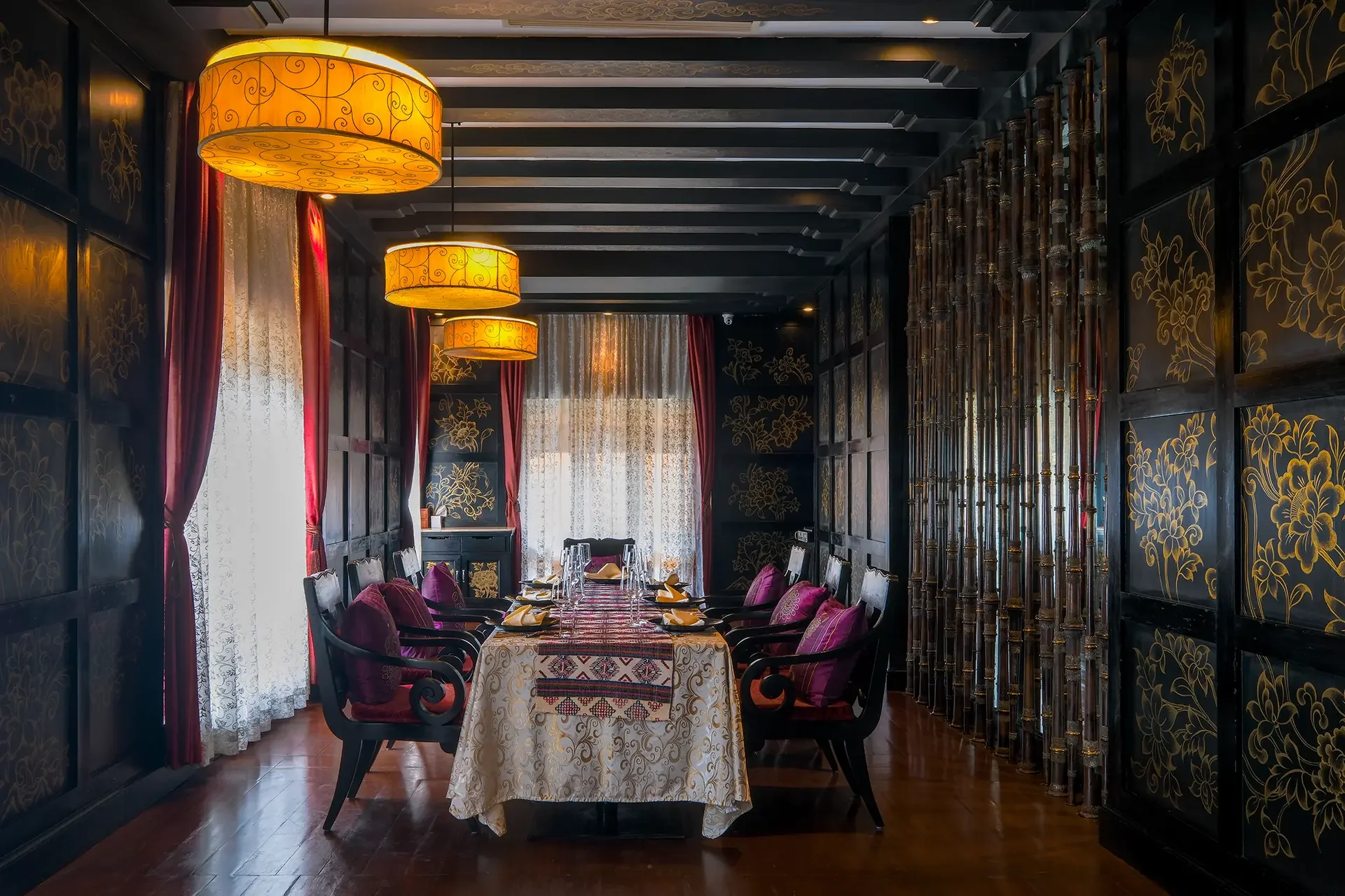 A dining room with a long table set for a meal, surrounded by dark wood chairs with purple cushions. The room has black walls with gold floral patterns, sheer white curtains, red velvet drapes, and ceiling lights with yellow shades. There are decorative bamboo poles on the right side of the room.