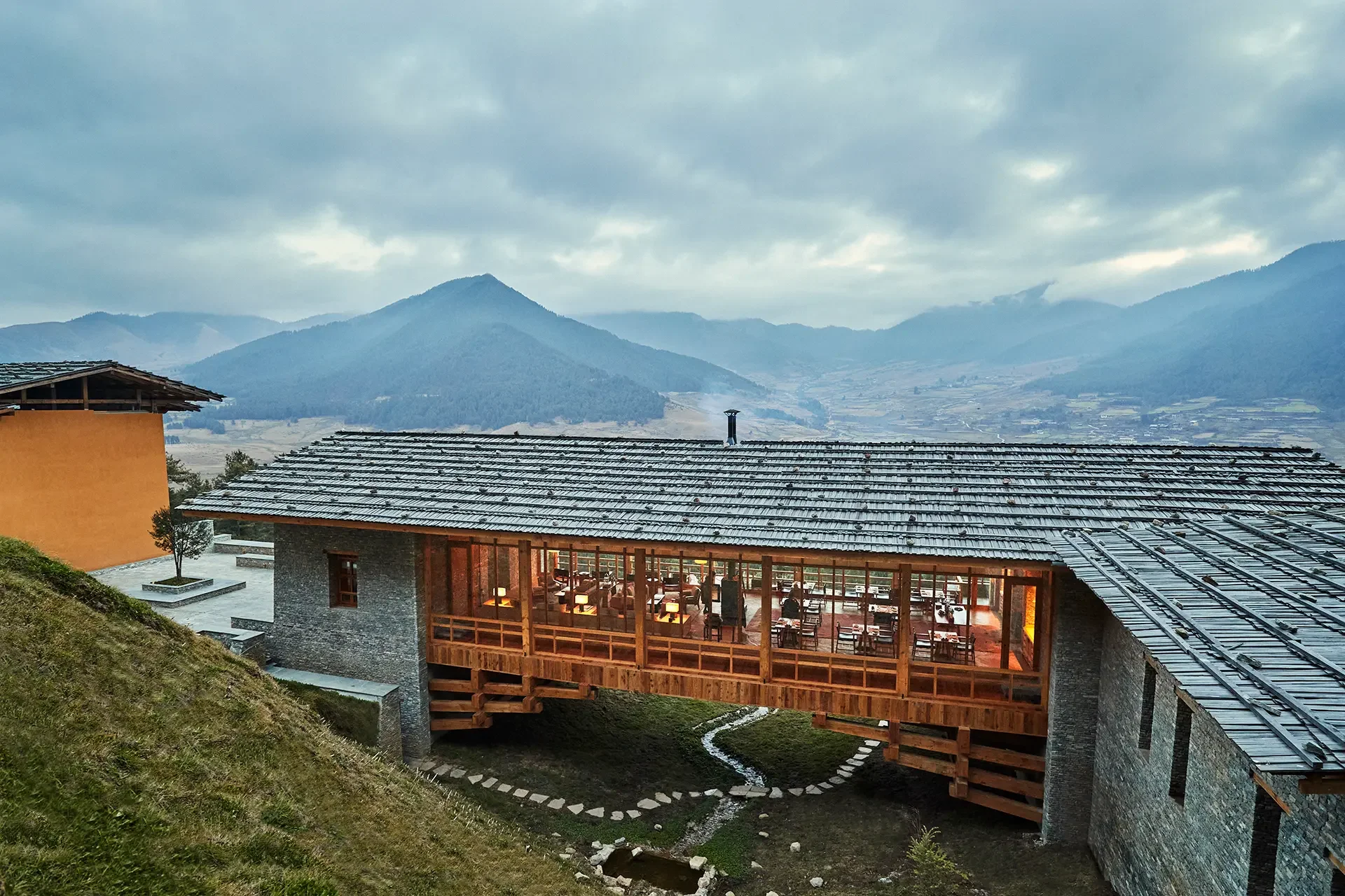 A scenic mountain view from a terrace with a restaurant or lounge area, showcasing a roofed outdoor seating space with wooden furniture, and mountain ranges in the distance under a cloudy sky.