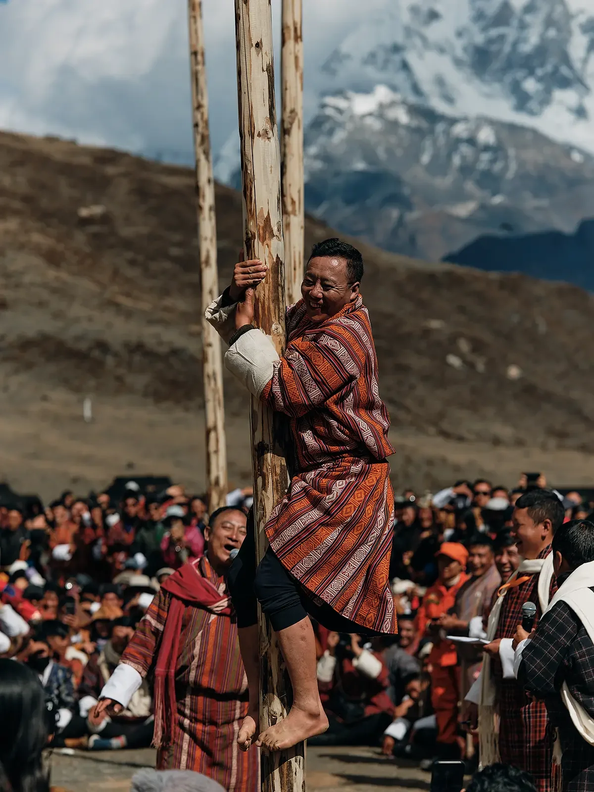 A man in traditional clothing is smiling and hugging a wooden pole while participating in a religious or cultural event. He is surrounded by a large crowd of people in similar attire, set against a mountainous landscape with snow-capped peaks in the 