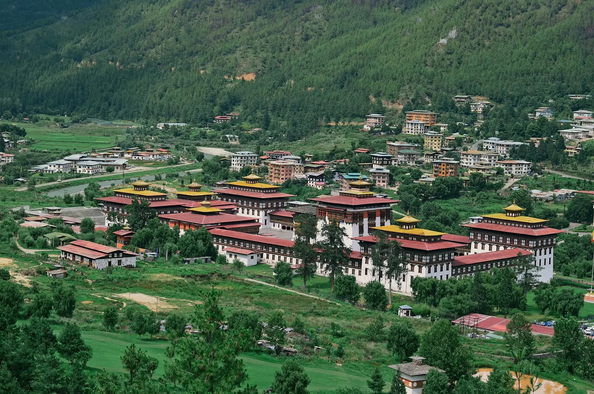 A scenic view of a lush green valley with multiple buildings, including traditional Tibetan-style structures with red and yellow roofs, situated among trees and fields, surrounded by forested hills.