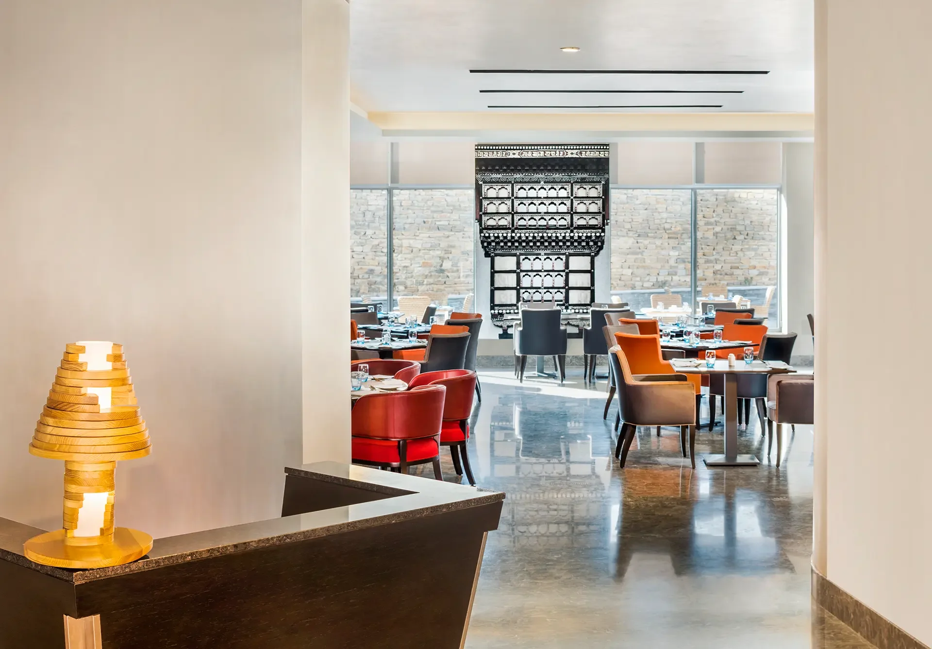 Interior view of a restaurant with tables and chairs, large windows, a stone wall, and decorative wall art. A wooden lamp is on a counter in the foreground.