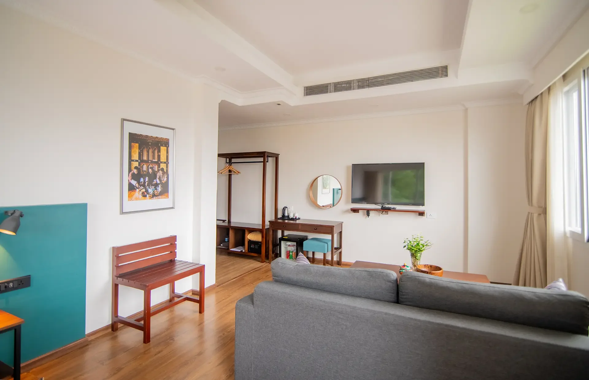 A bright hotel room with wooden floors, a gray sofa, a wall-mounted TV, a round wall mirror, a desk with a chair, a small table with a plant, a framed picture on the wall, and large windows with curtains.