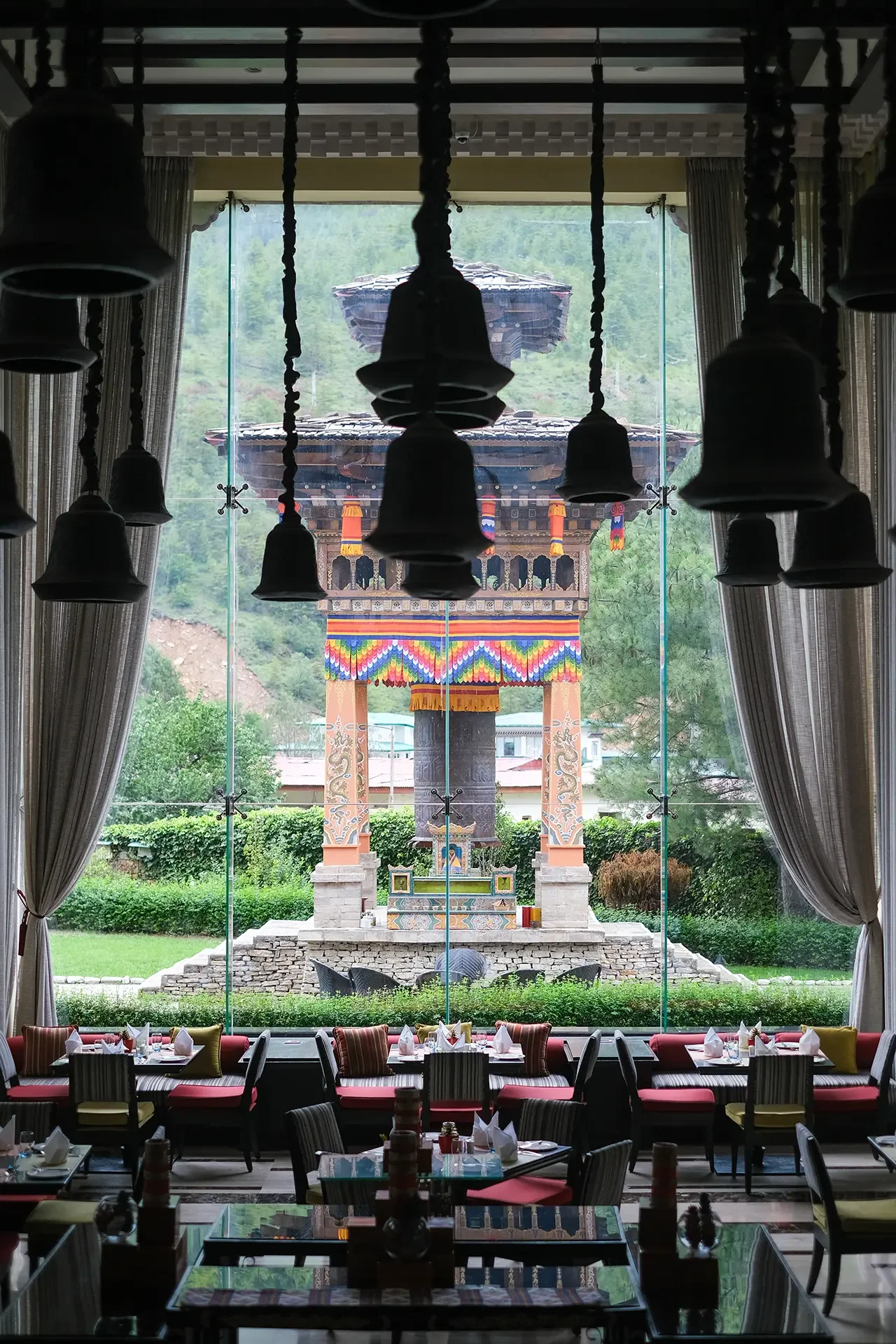 Interior view of a restaurant with large glass window showing a traditional temple gate outside. The temple has a multi-tiered roof and colorful decorations, surrounded by greenery. Inside, there's a dining area with tables, chairs, and hanging lamps.