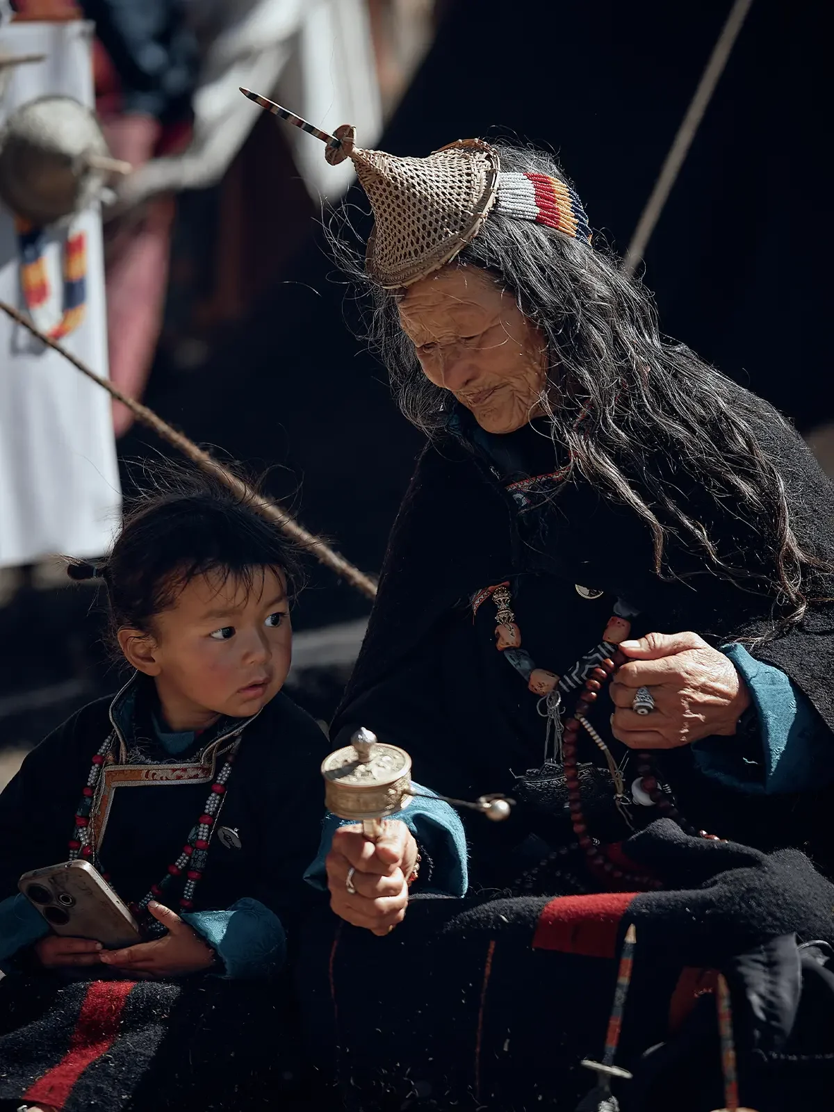 An older woman and a young girl dressed in traditional clothing, sitting together outdoors. The woman wears a headdress with a long feather and beaded accessories, while the girl holds a small musical instrument and a smartphone.