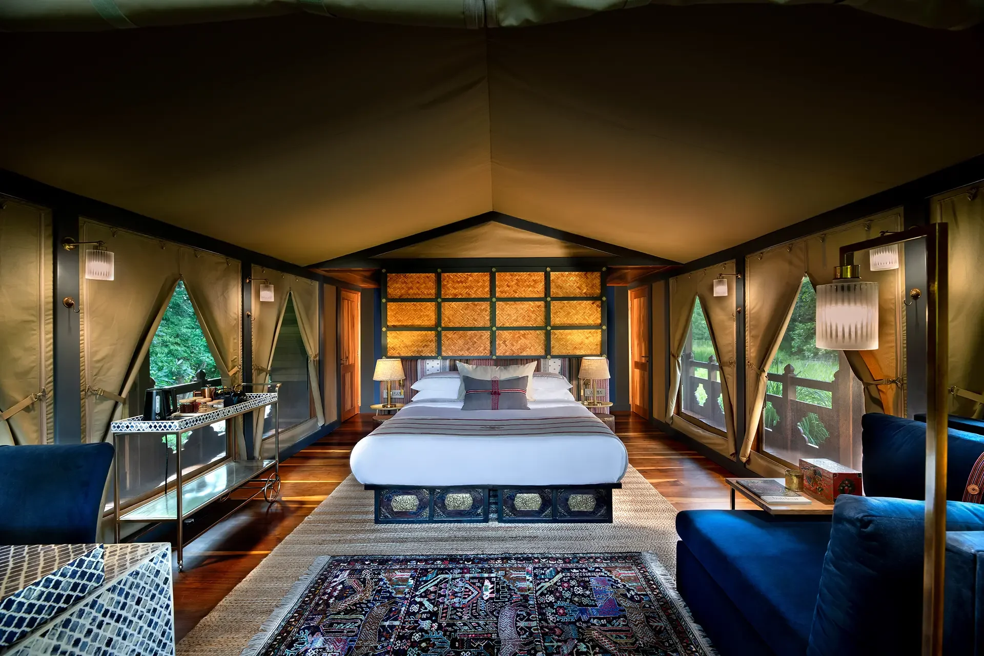 A bedroom with large windows draped with beige curtains, a bed with white linens and multiple pillows, two bedside lamps, colorful rugs, and a view of green trees outside.