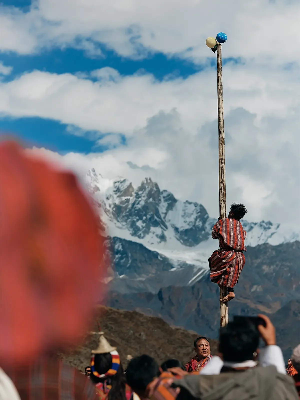 A boy in traditional clothing climbs a tall wooden pole topped with colorful balls against a mountainous background with snow-capped peaks under a cloudy sky, with people gathered around watching.