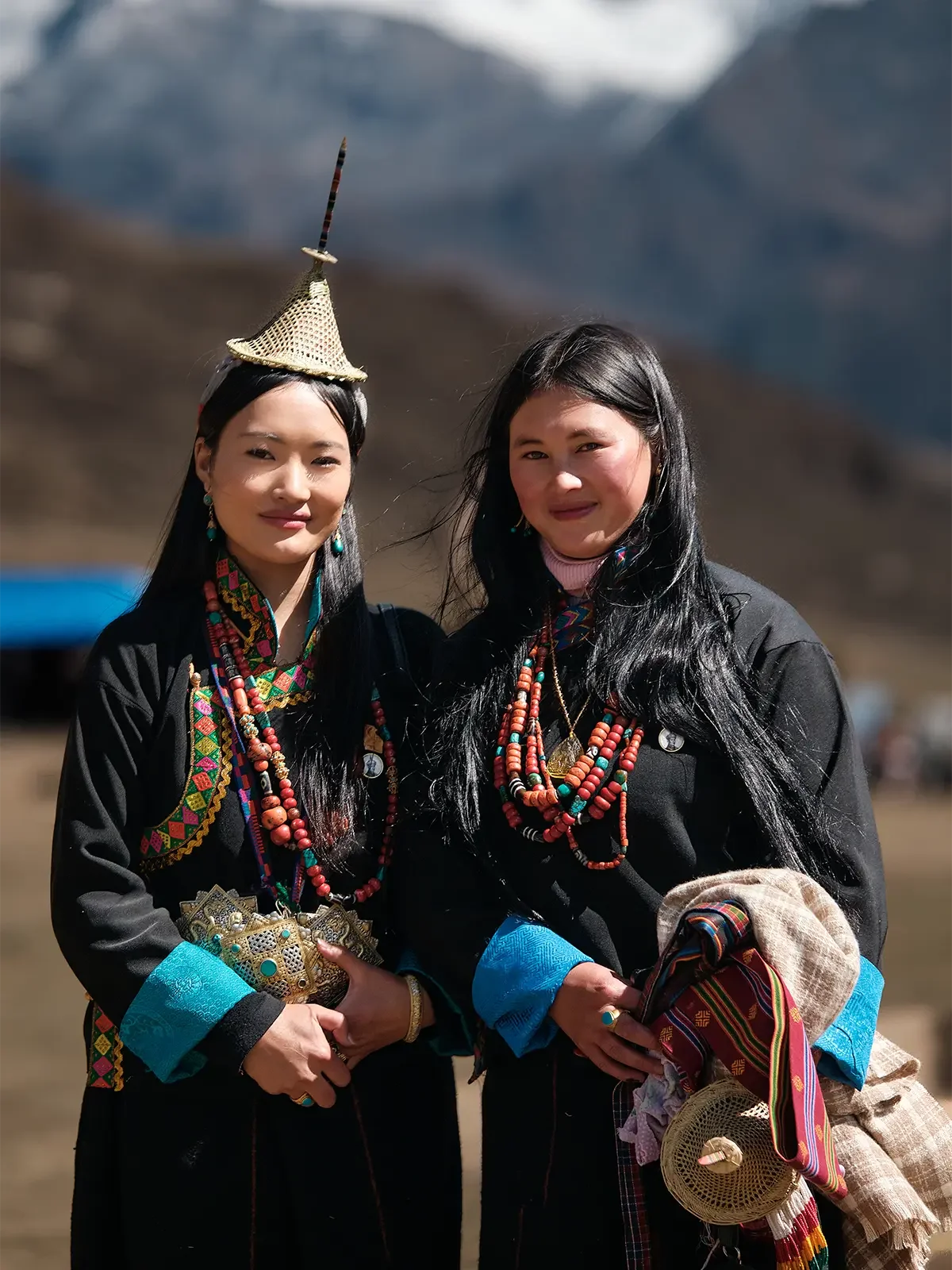 Two women wearing traditional clothing and jewelry, standing outdoors with mountains in the background.