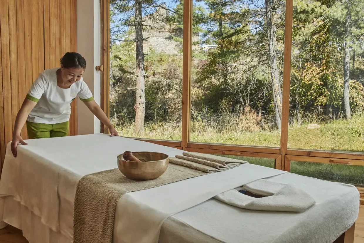 A woman in a spa or massage room preparing a massage bed with a white sheet, with a large window showing trees and greenery outside.