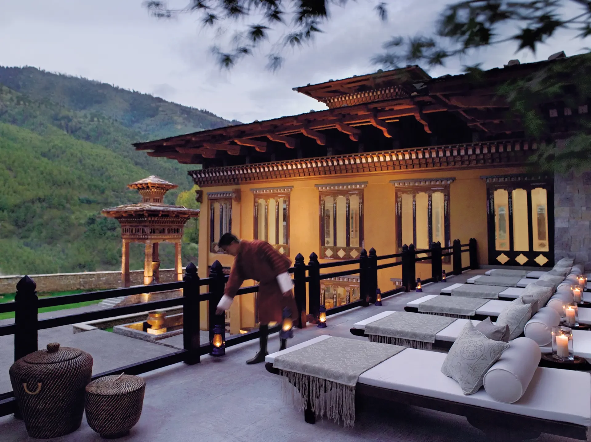 A scenic outdoor terrace with decorative lanterns, cushioned seating, and a man setting up cushions, with traditional wooden architecture and lush green mountains in the background.