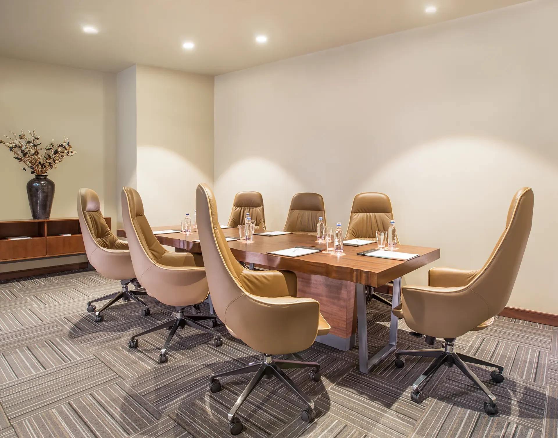 A conference room with a long wooden table and beige leather chairs around it. On the table are water bottles, glasses, and notepads. The room has a light-colored wall and carpeted floor. There is a tall vase with decorative branches on a side table.
