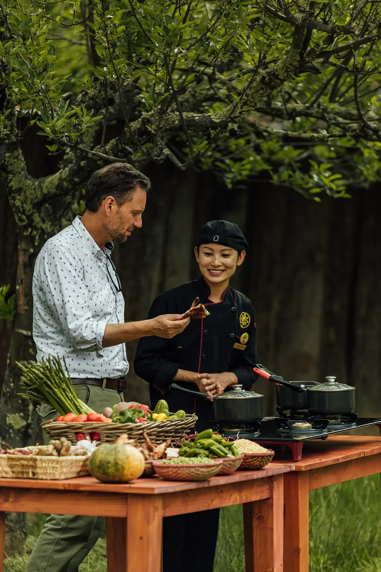 A man and a woman in chef attire standing behind a table of fresh vegetables and cooking pots outdoors, with trees and green foliage in the background.