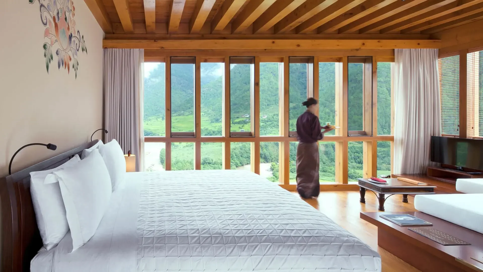 A hotel room featuring a large bed with white bedding and pillows, a nightstand and reading lamps, a flat-screen TV on a wooden stand, and a woman in traditional attire standing by a window with a view of green mountains and fields.