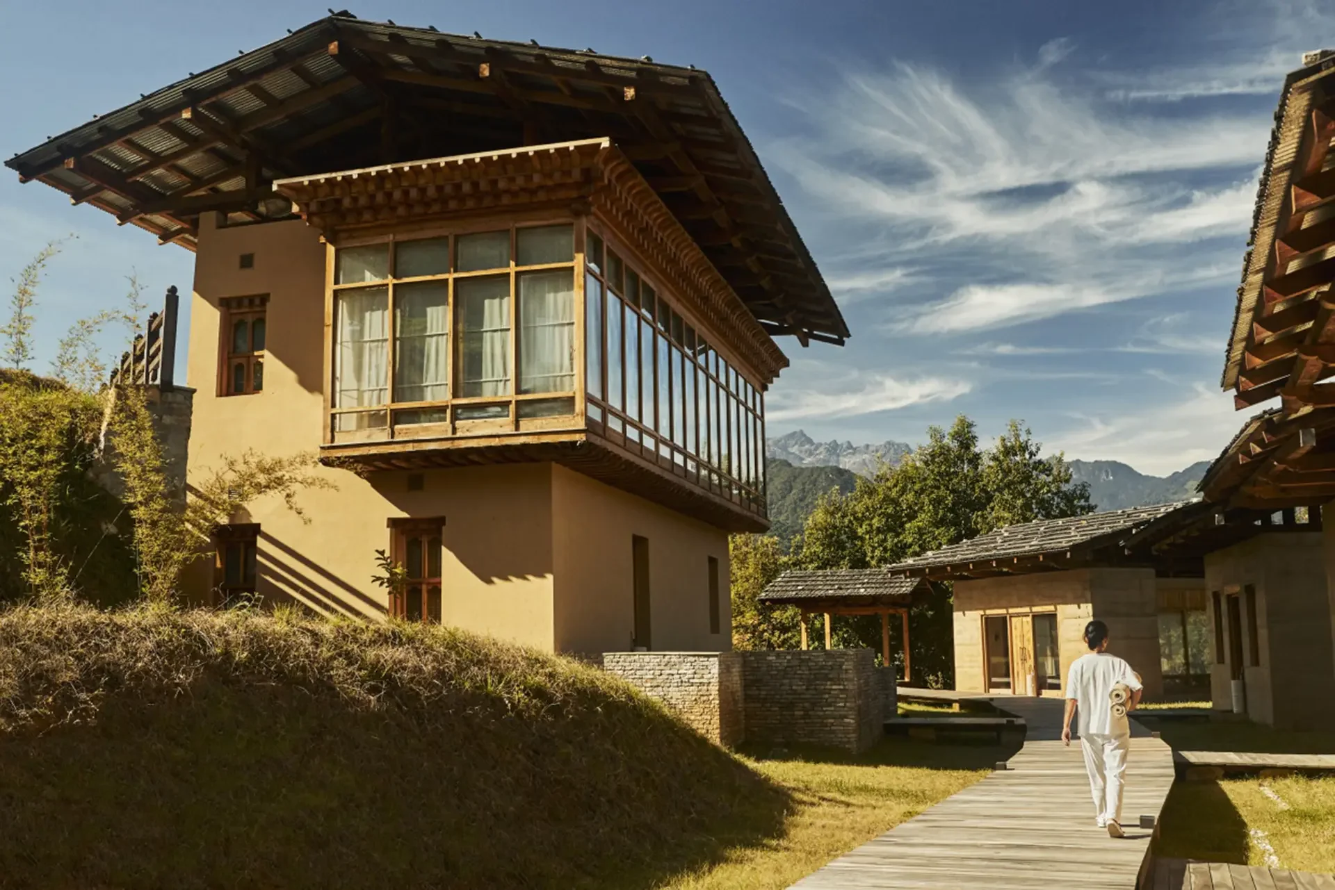 A person walking on a wooden pathway towards a traditional-style house with large windows, set in a mountainous landscape with clear skies and trees.